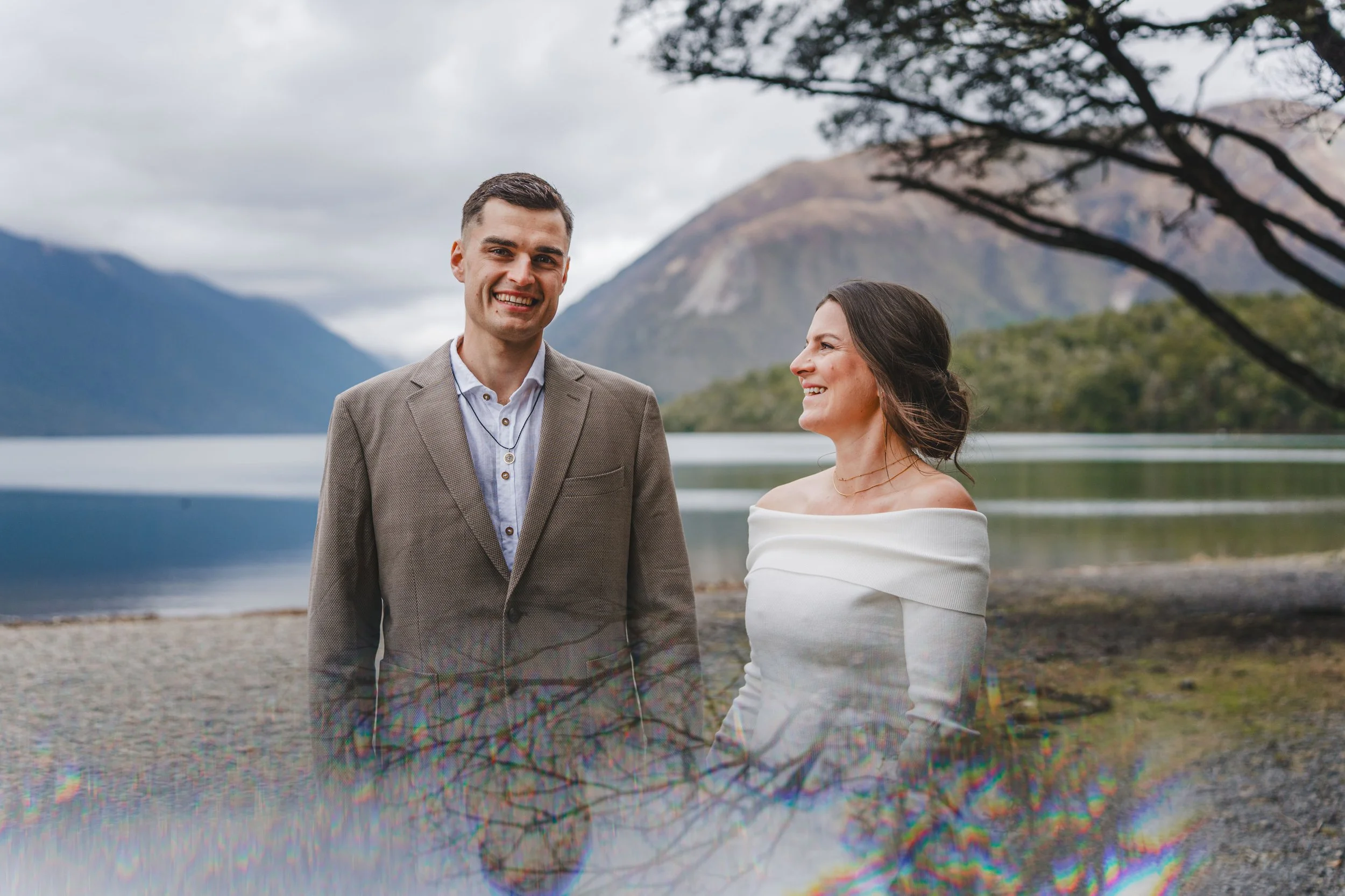 A smiling man and woman holding hands by a lake with mountains in the background, the man is wearing a beige suit jacket and a dress shirt, the woman is wearing an off-shoulder white dress.