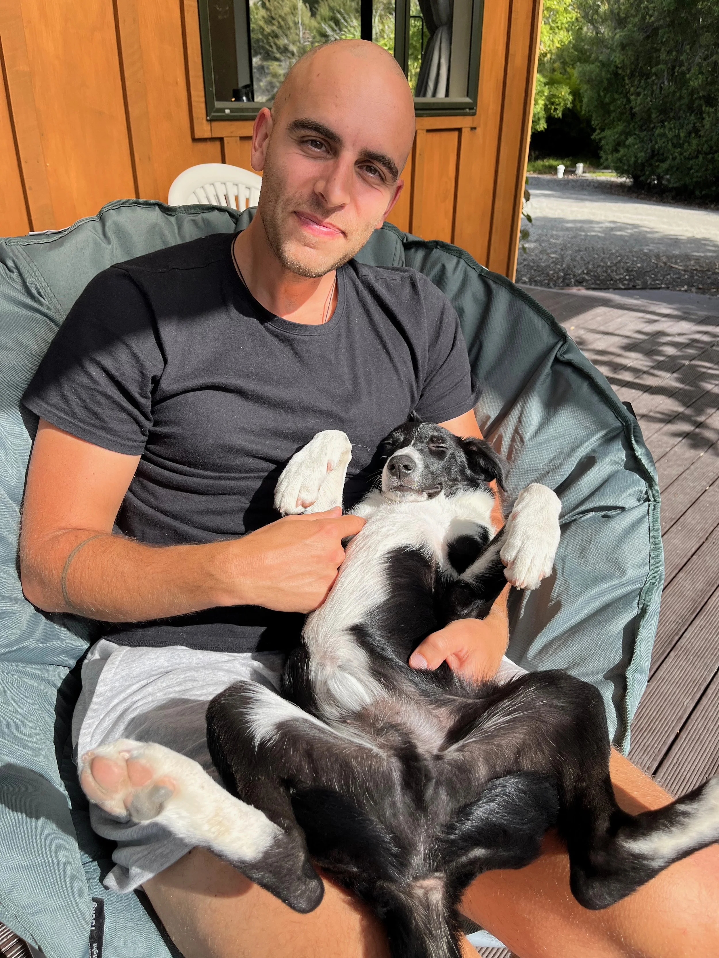 A man in a black t-shirt sitting outside on a cushioned chair, holding a black and white puppy on his lap, as the puppy lies on its back with eyes closed. The background shows wooden siding and part of a deck.