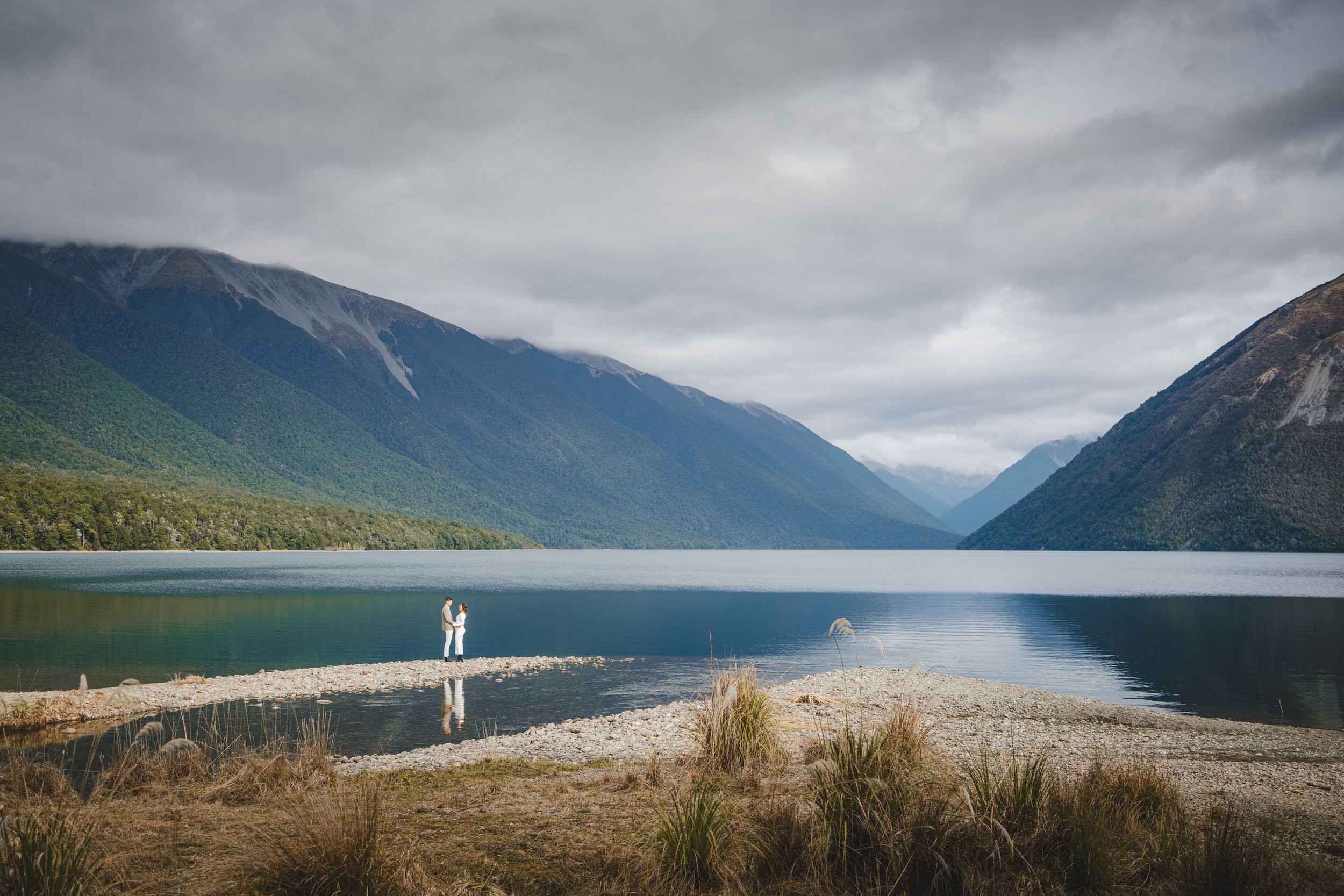 A couple standing on a small strip of land at the edge of a lake, surrounded by mountains and overcast sky.