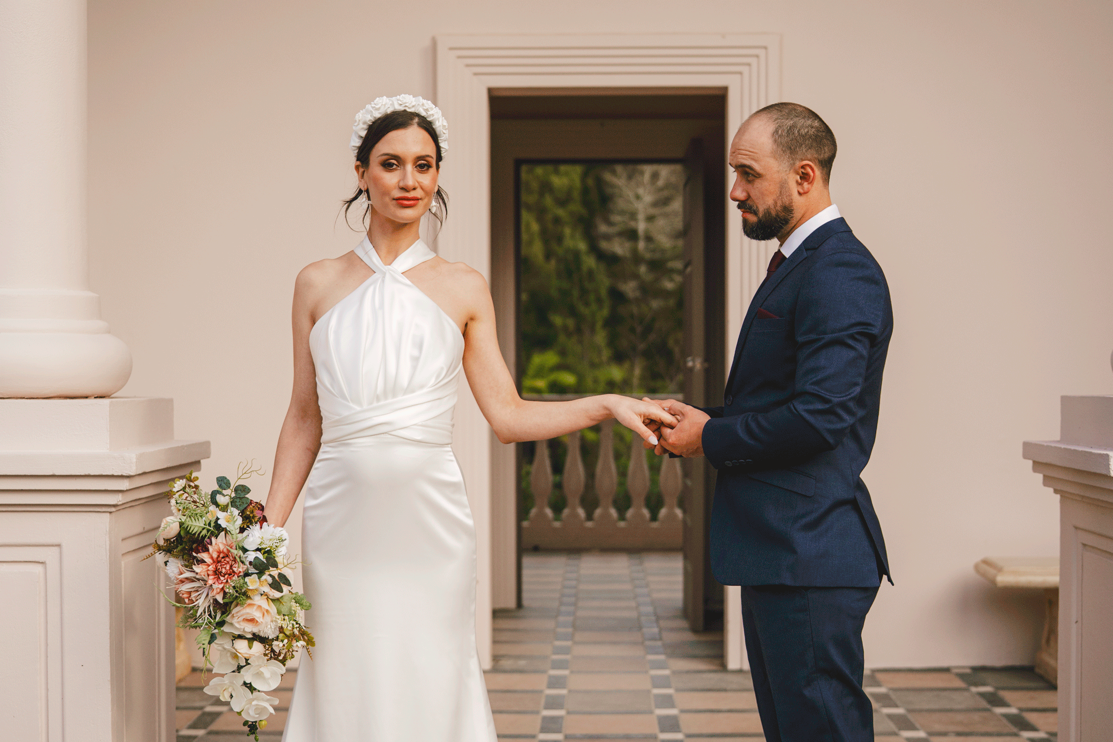 A bride in a white halter wedding dress holding a bouquet, and a groom in a dark blue suit, holding hands during their wedding vows inside a building with a doorway and a scenic outdoor background.