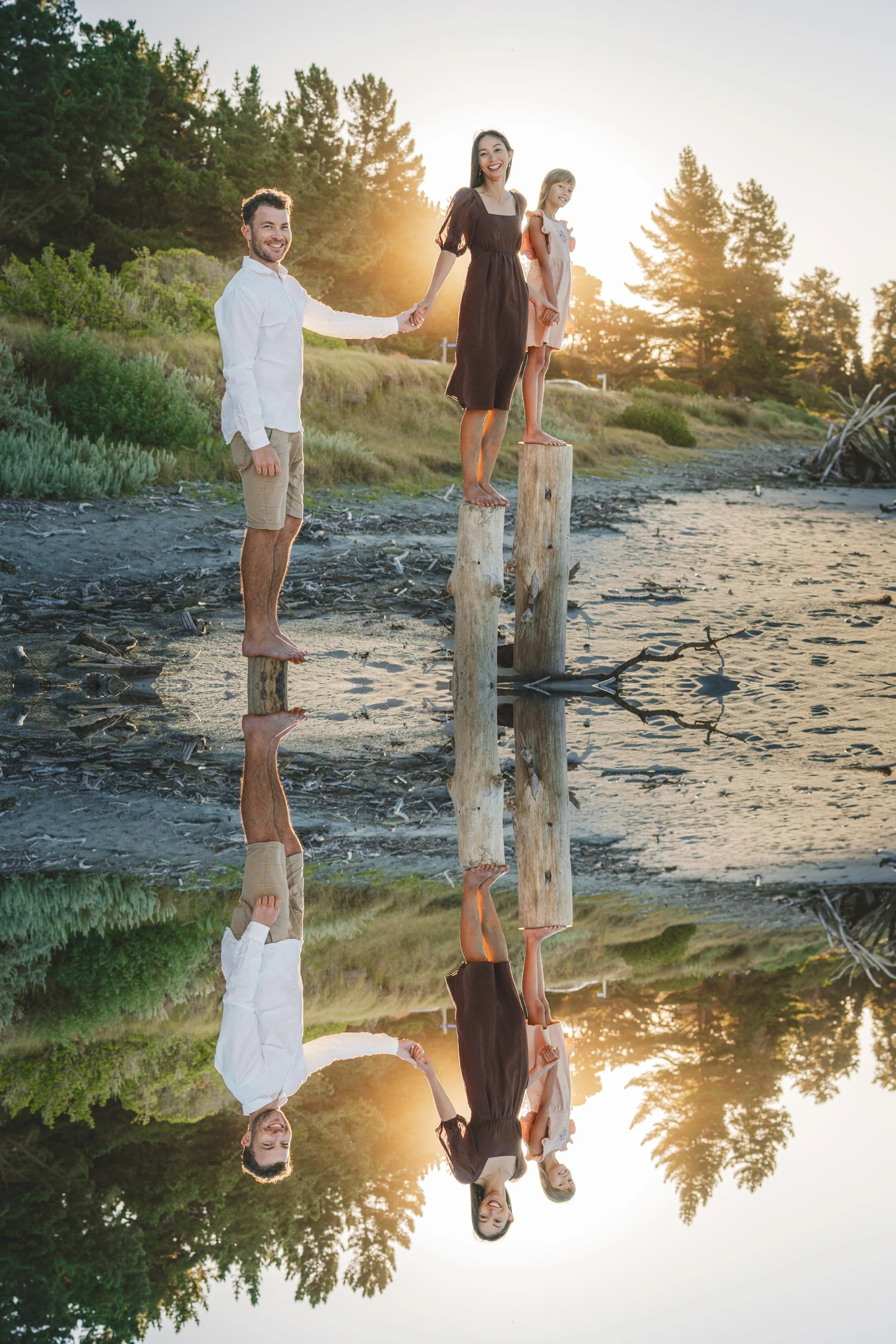 A family of three standing on wooden posts at the beach during sunset, holding hands, with their reflection visible in the water.