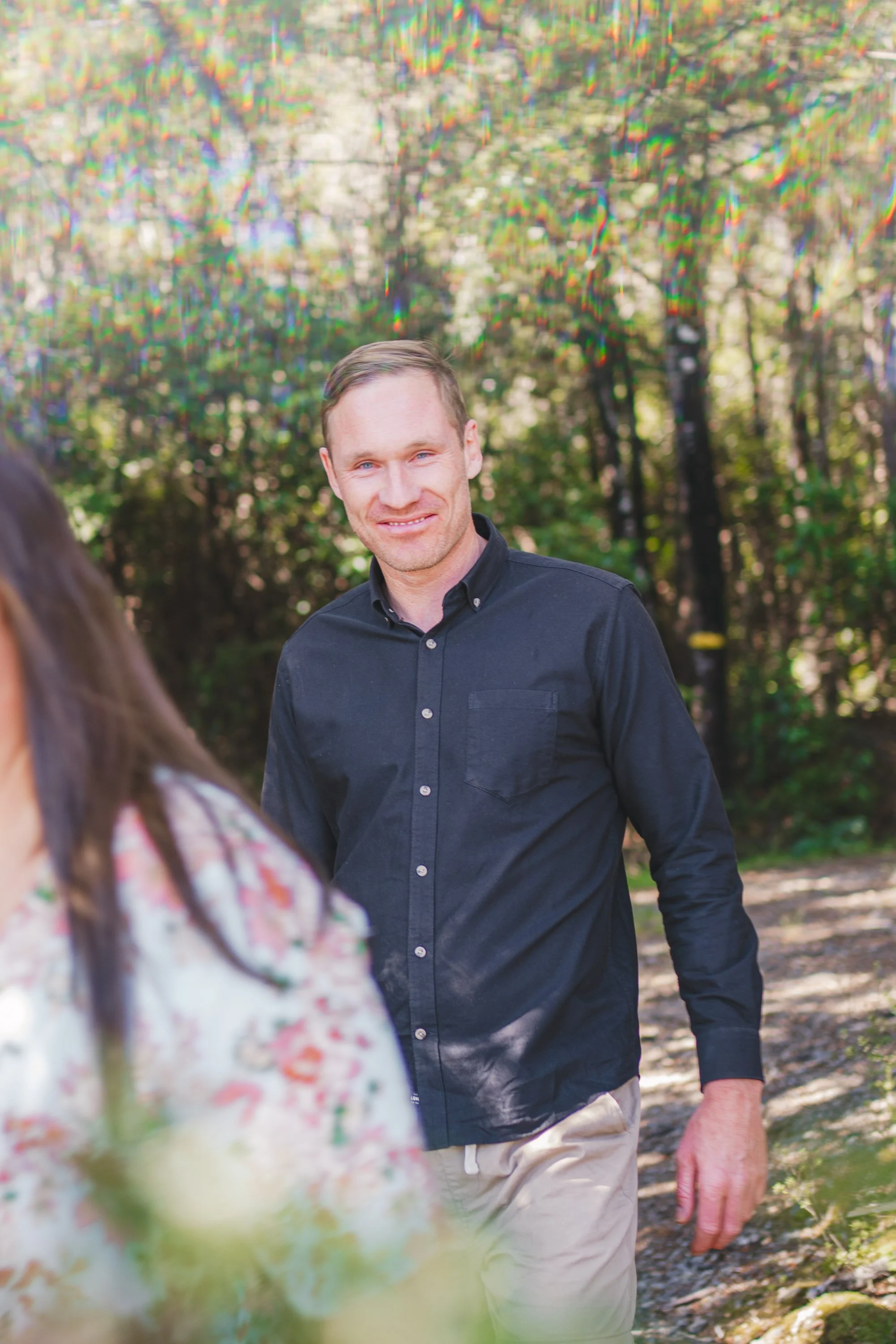 A man with short light brown hair wearing a dark button-up shirt and khaki pants, smiling while walking outdoors in a wooded area with sunlight filtering through the trees.