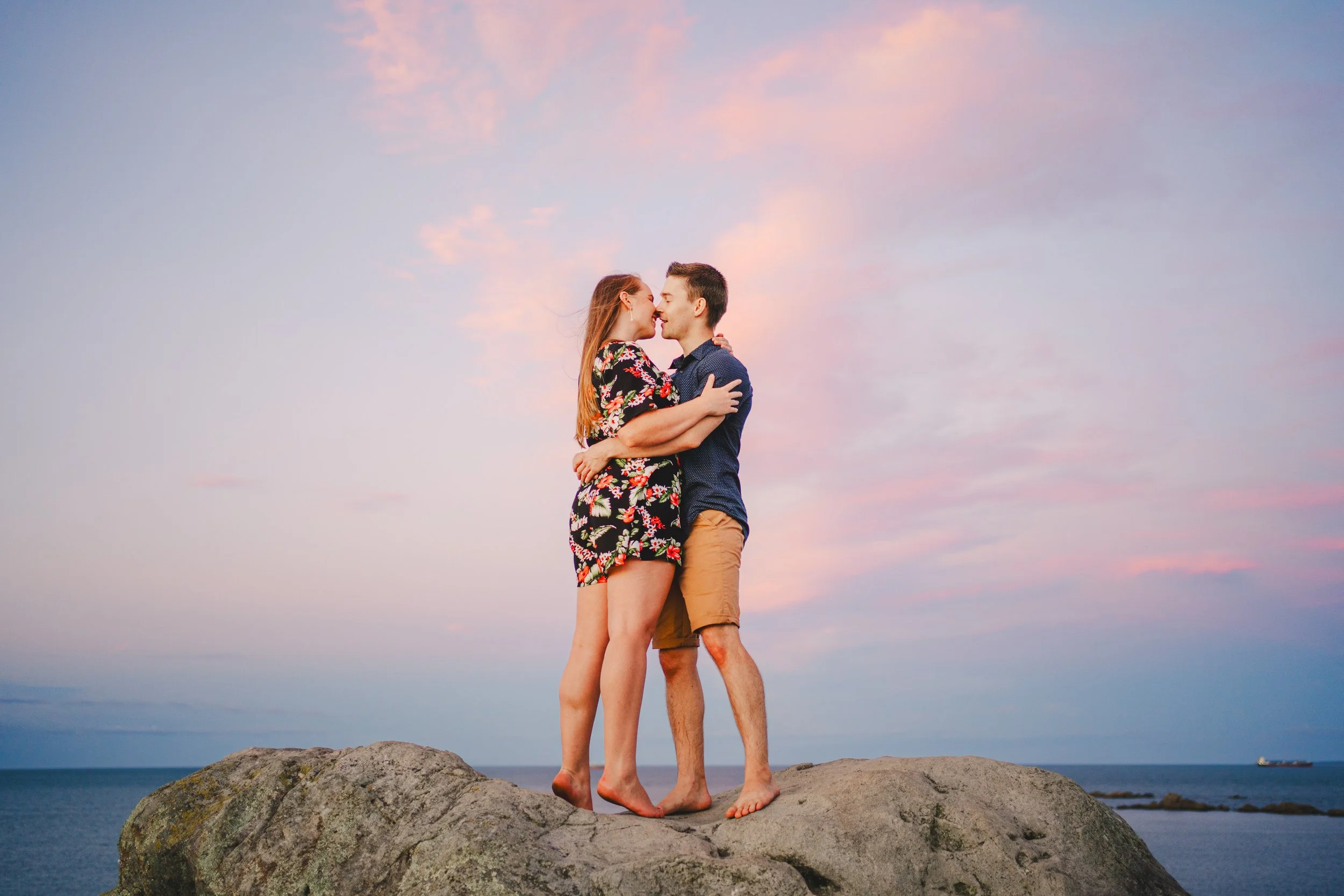 A couple standing on a large rock at the beach, embracing and about to kiss at sunset, with a pastel-colored sky and water in the background.