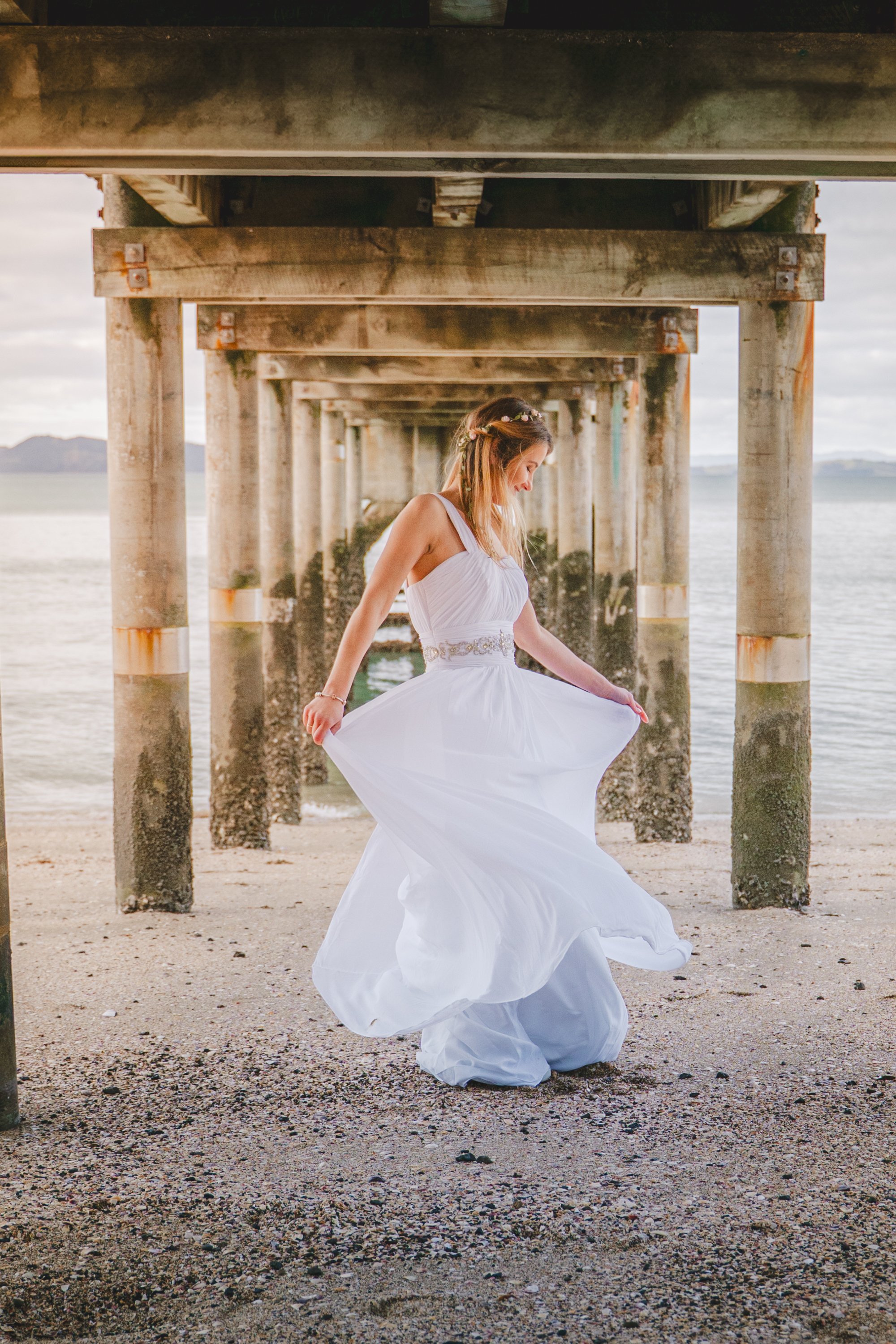 A young woman in a white dress standing under a weathered pier on a beach.