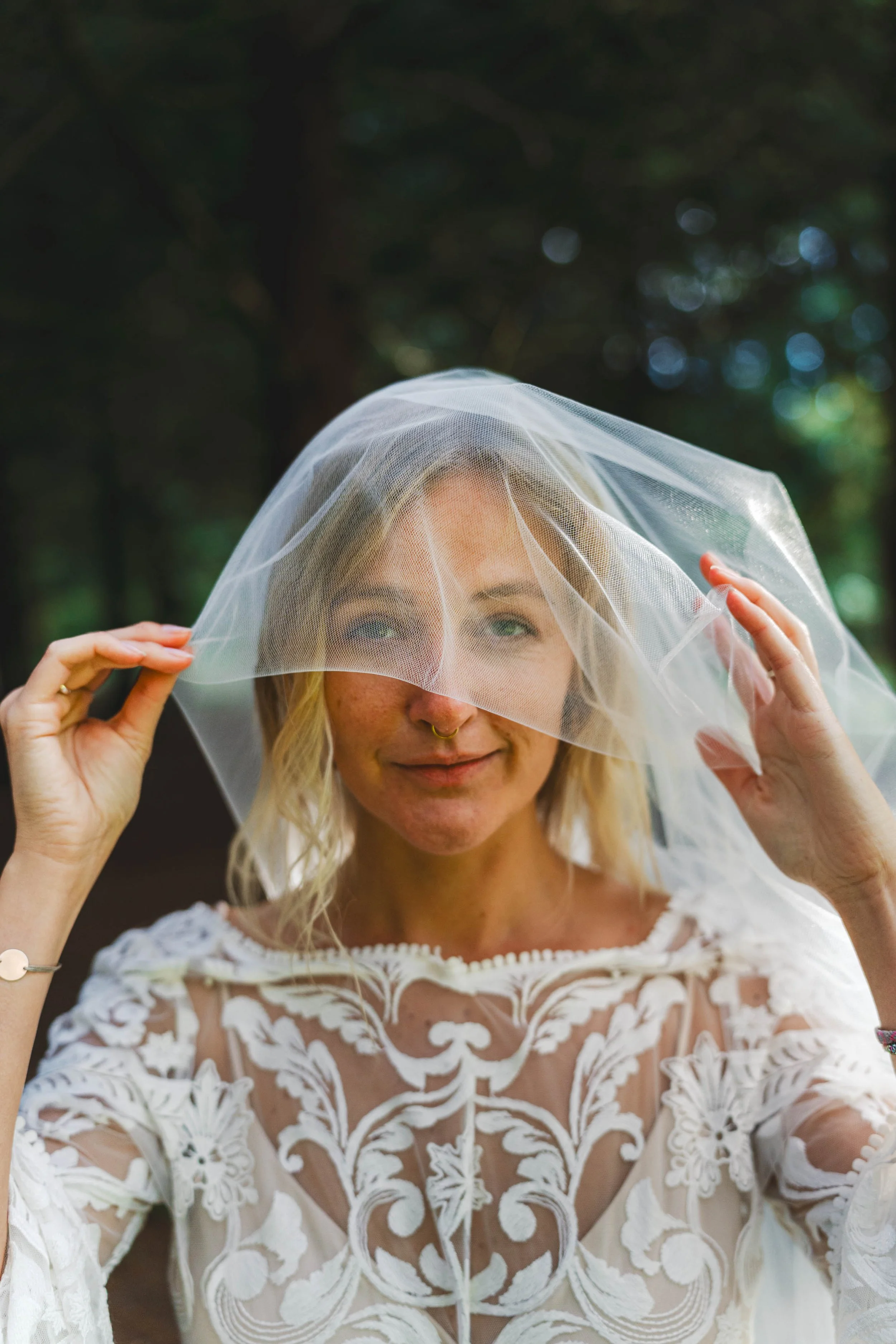 A woman with blonde hair, wearing a white embroidered dress, holds a sheer veil over her face, smiling softly with a forest background.