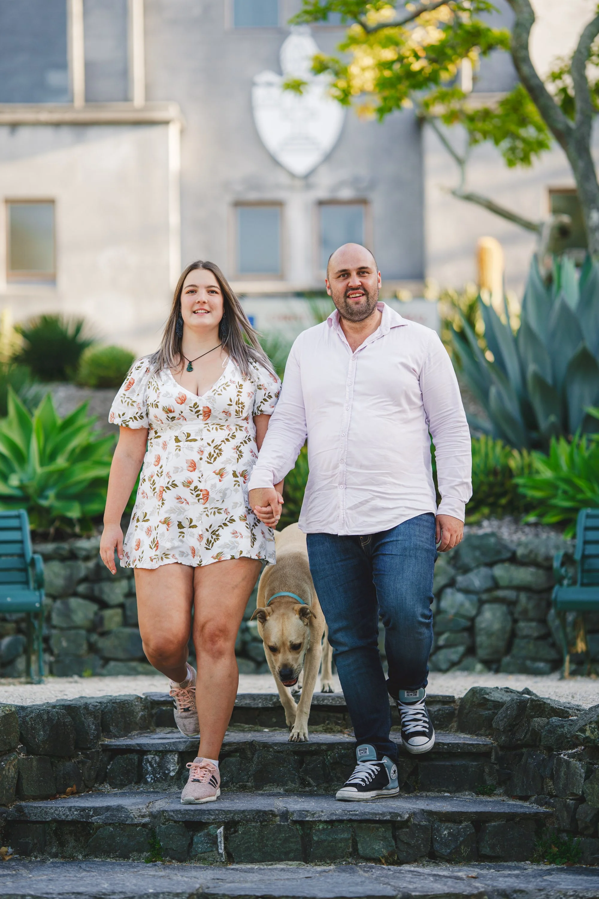 A man and woman walking down stone steps holding hands with a dog, in a garden with green plants and a building in the background.