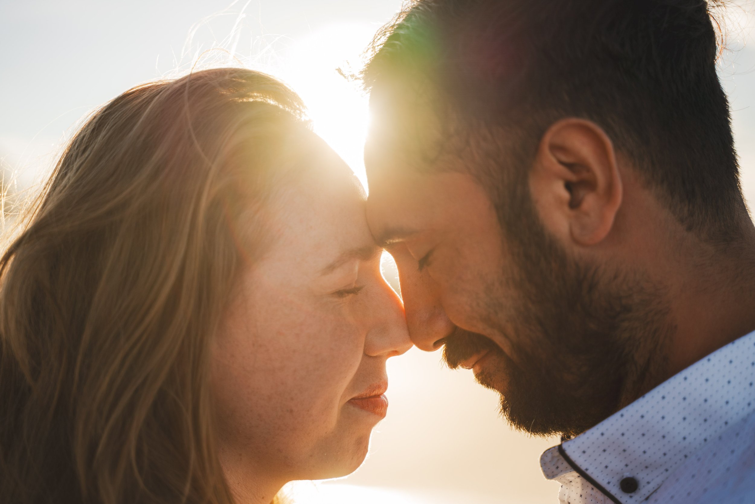 A close-up of a couple with their foreheads pressed together and eyes closed, sunlight shining behind them.