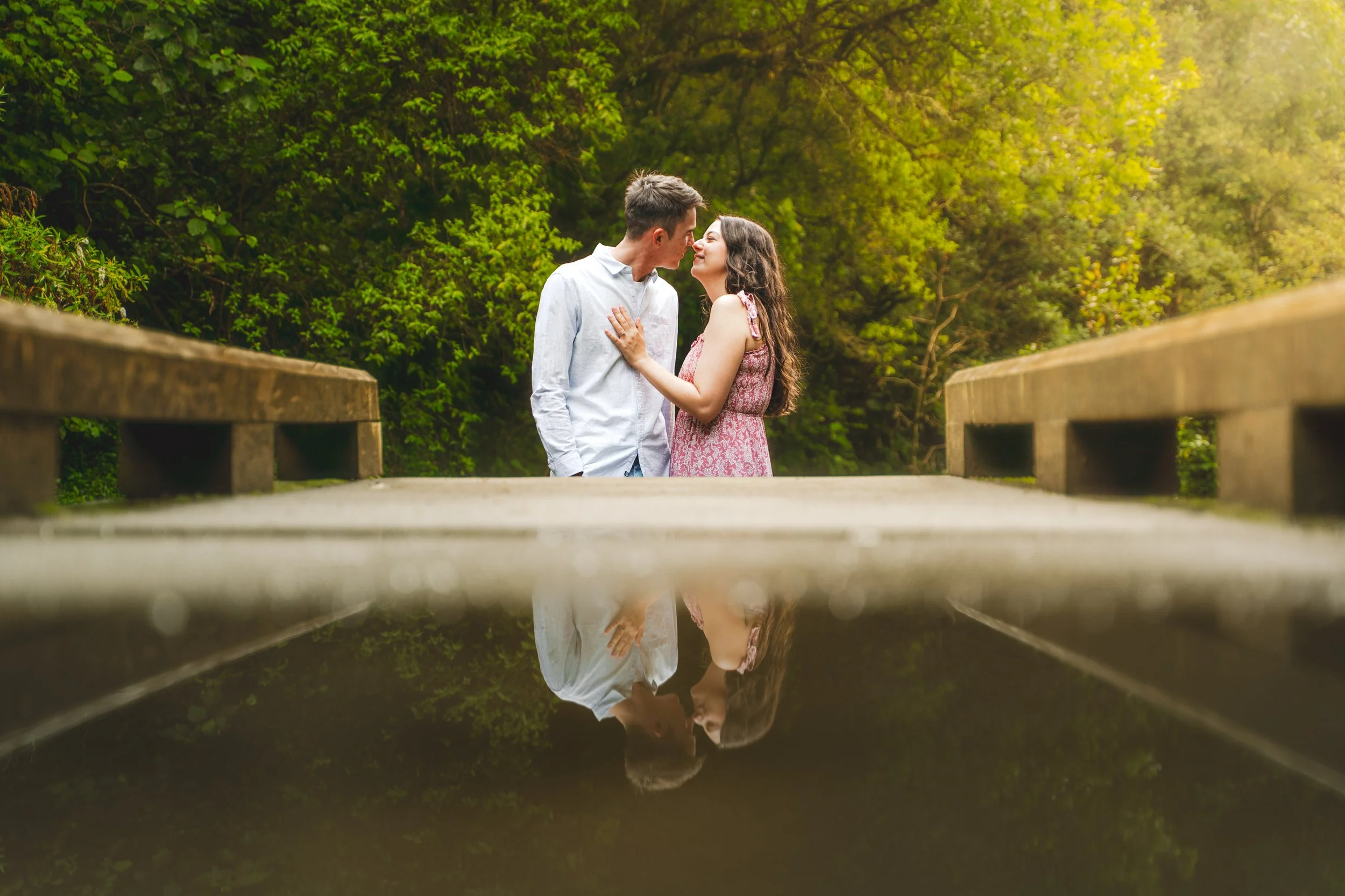 A man and woman standing on a bridge in a lush green park, leaning towards each other, about to kiss, with their reflection visible in a puddle beneath them.