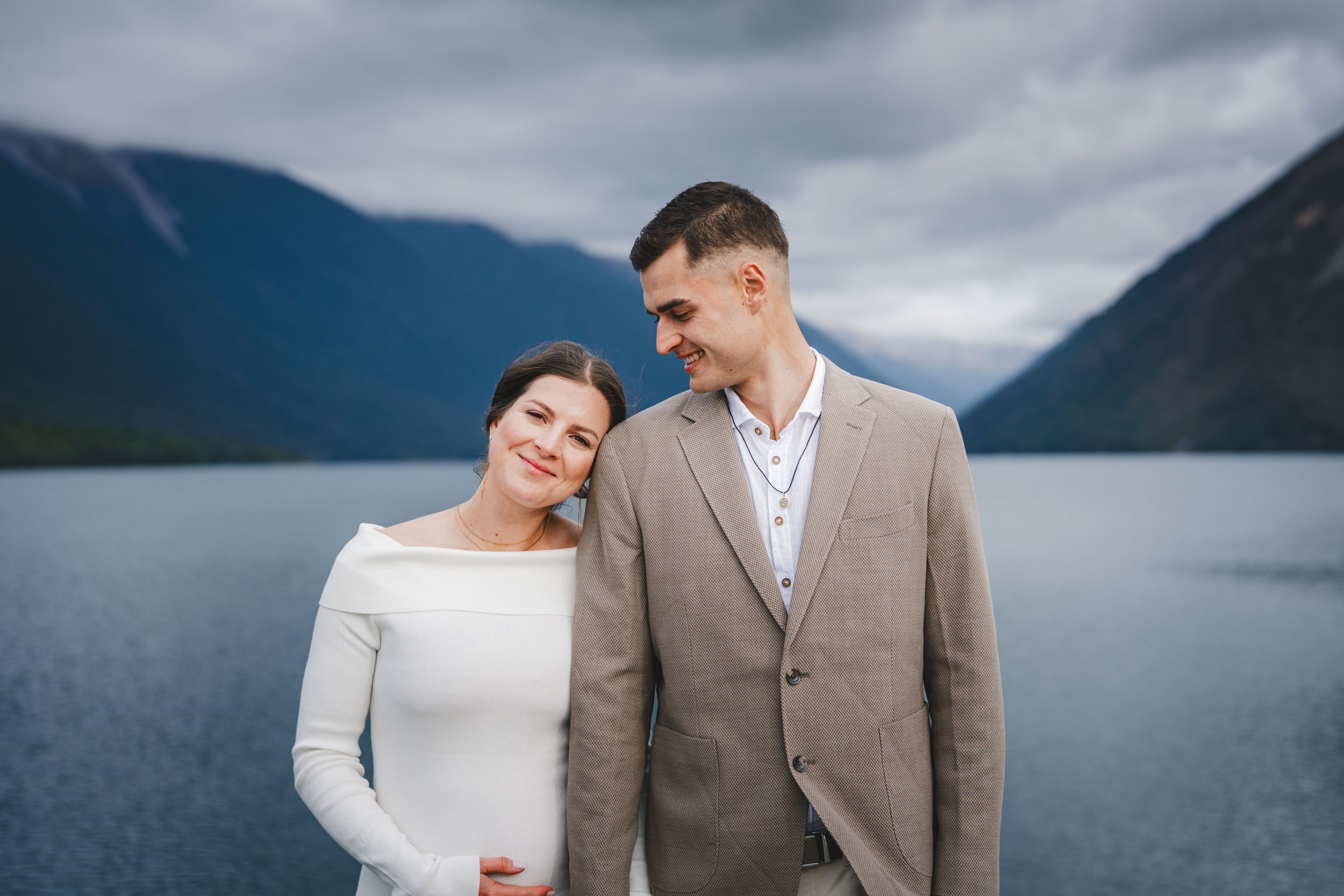 A couple standing close to each other outdoors near a body of water with mountains and cloudy sky in the background. The woman, wearing a white dress, is resting her head on the man's shoulder and smiling. The man, dressed in a beige suit, is looking