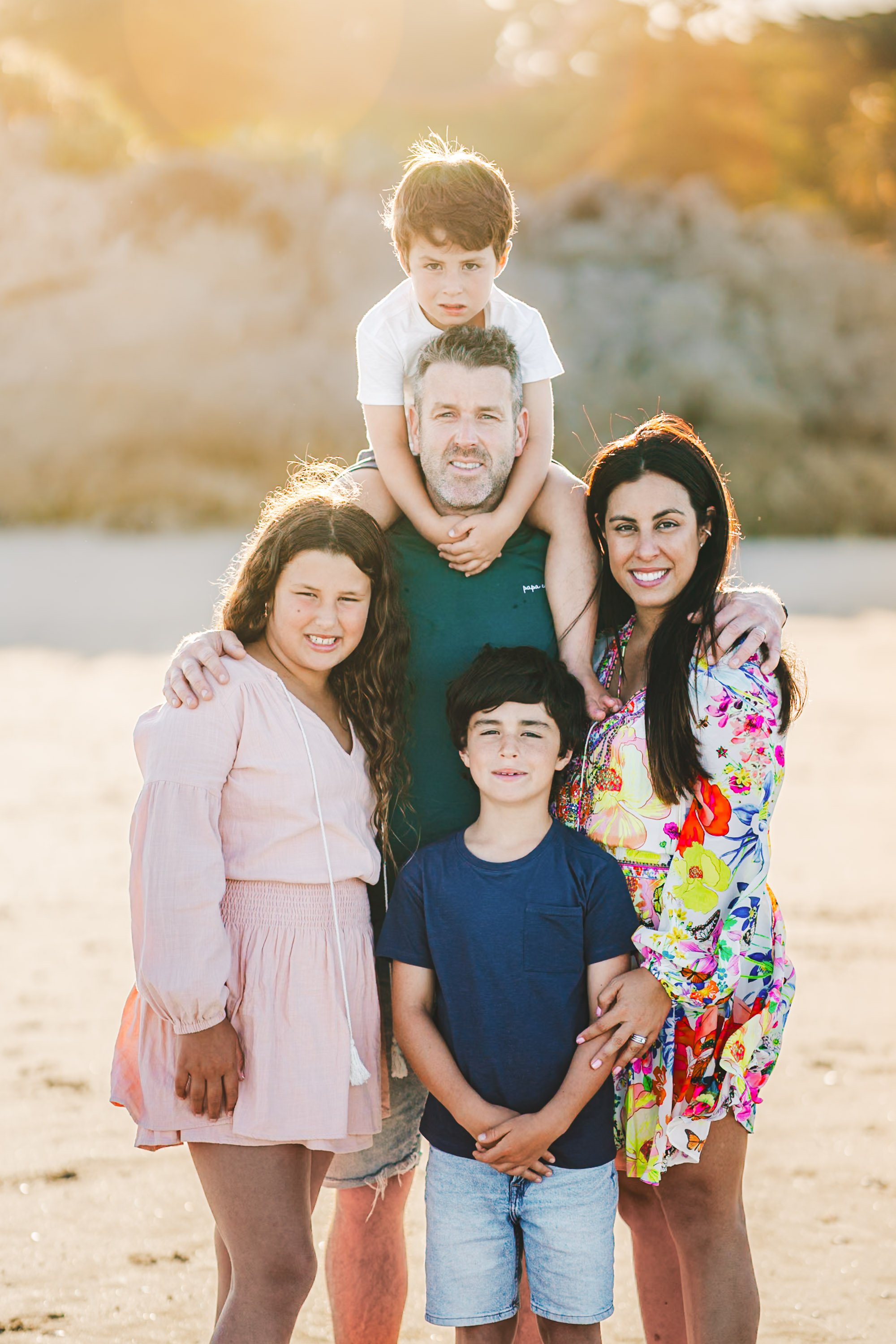 A family of five posing outdoors on a sunny day, with a rocky background. The family includes a man, woman, and three children, all smiling at the camera.