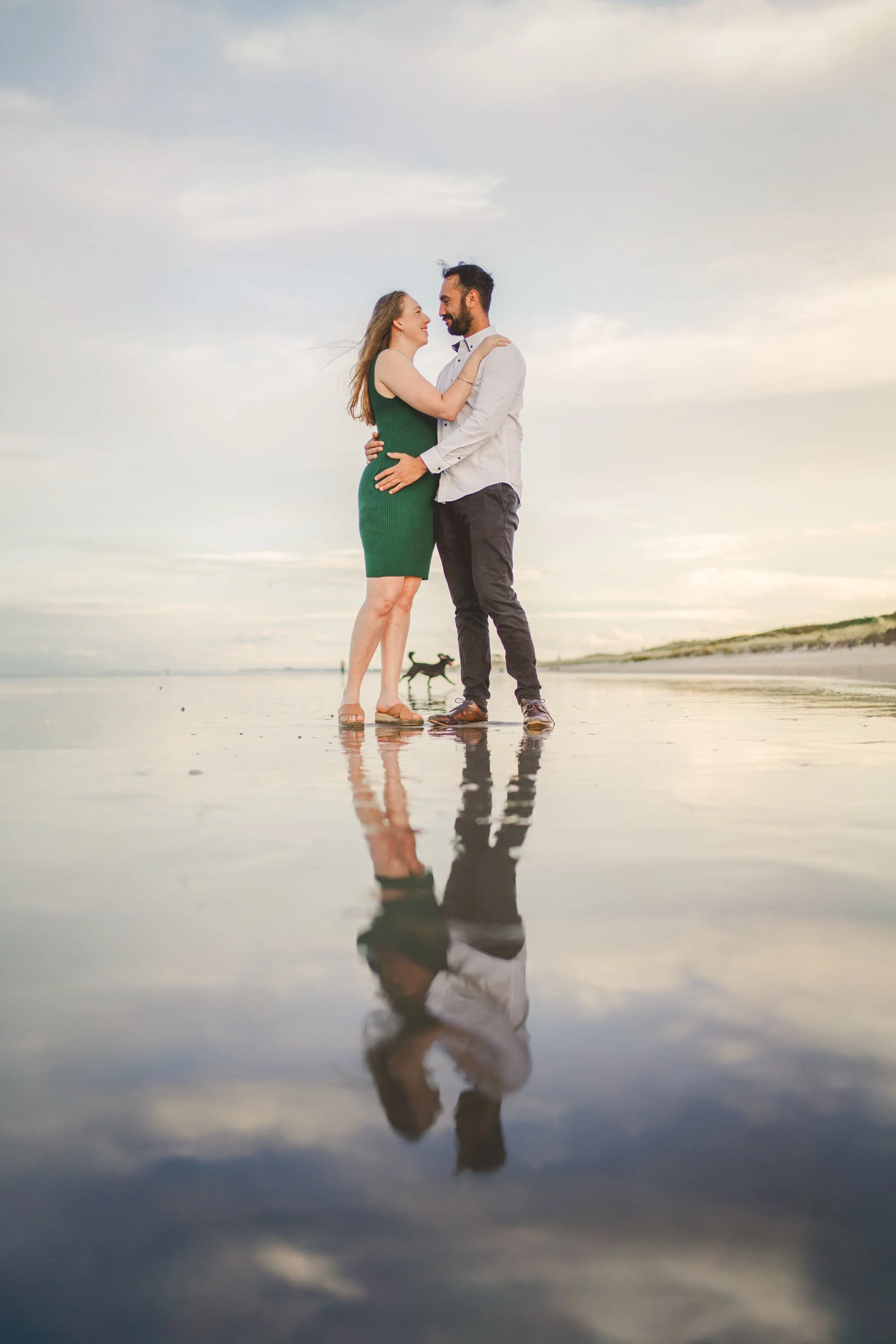 A couple embracing on a beach with their reflection visible in the water, and a dog running in the background.