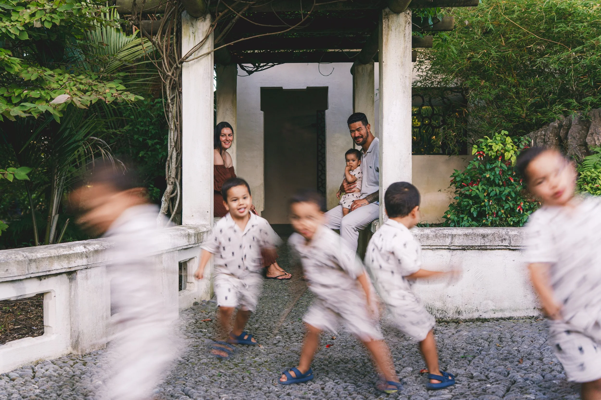 Family and children playing outside near a white garden structure with greenery, while a man, woman, and baby sit and smile in the background.