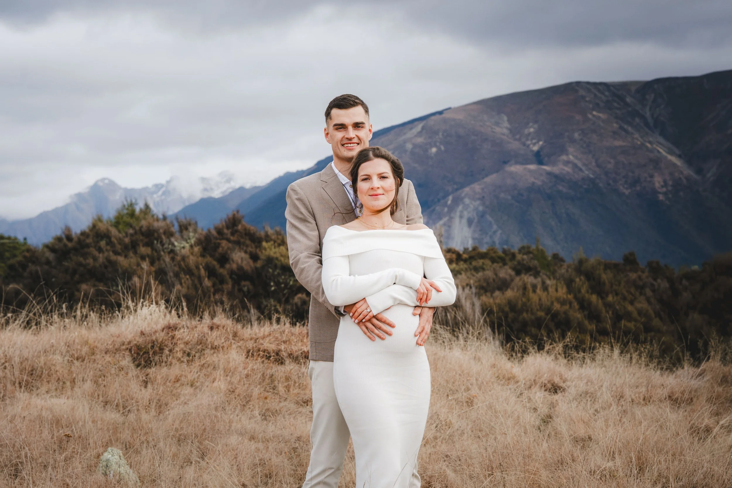 A couple stands outdoors in a mountainous landscape during daytime, dressed formally with the man in a beige blazer and the woman in a white dress, both smiling and embracing.