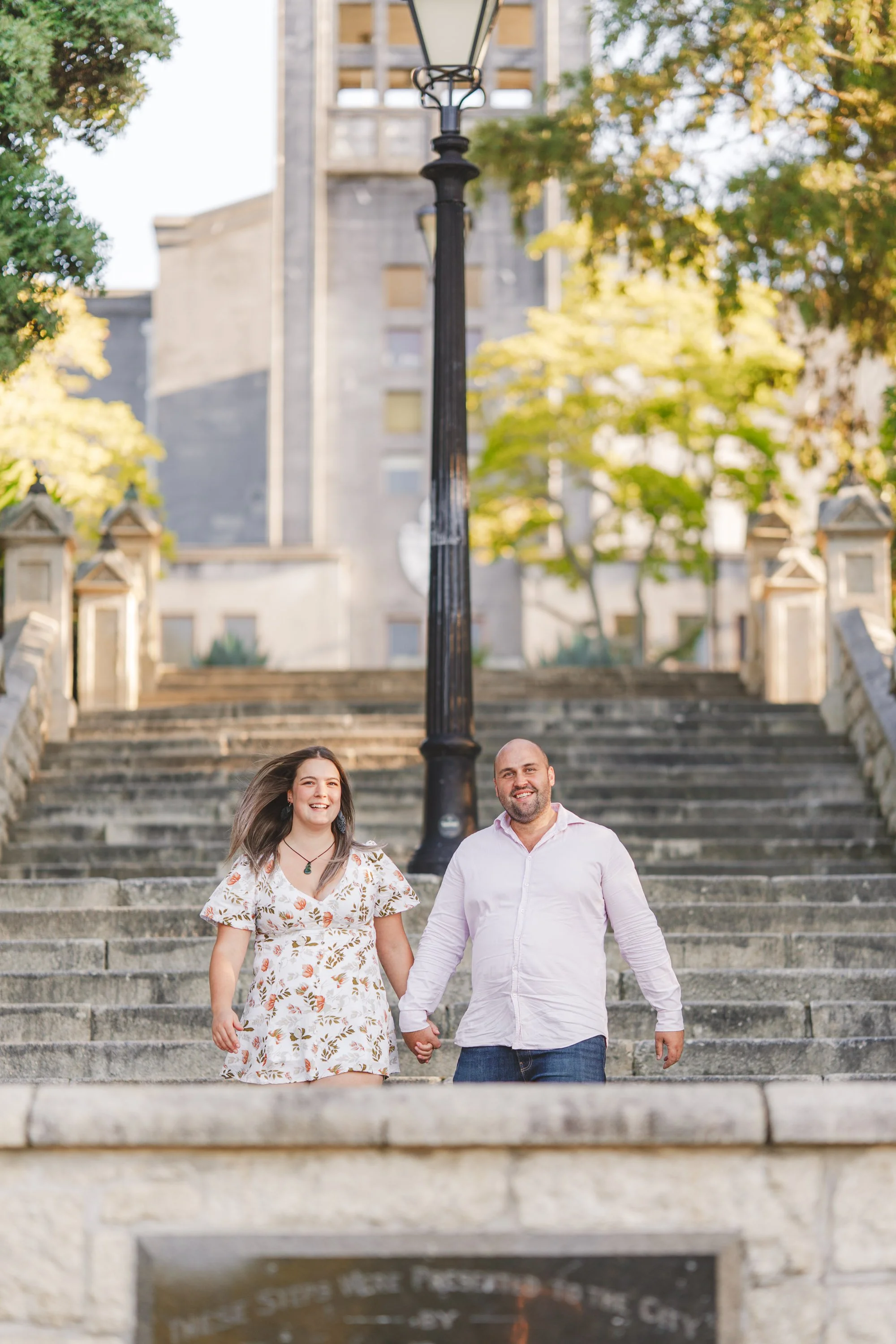 A smiling couple holding hands and walking down stone stairs outdoors in a park setting, with trees and a historic building in the background.