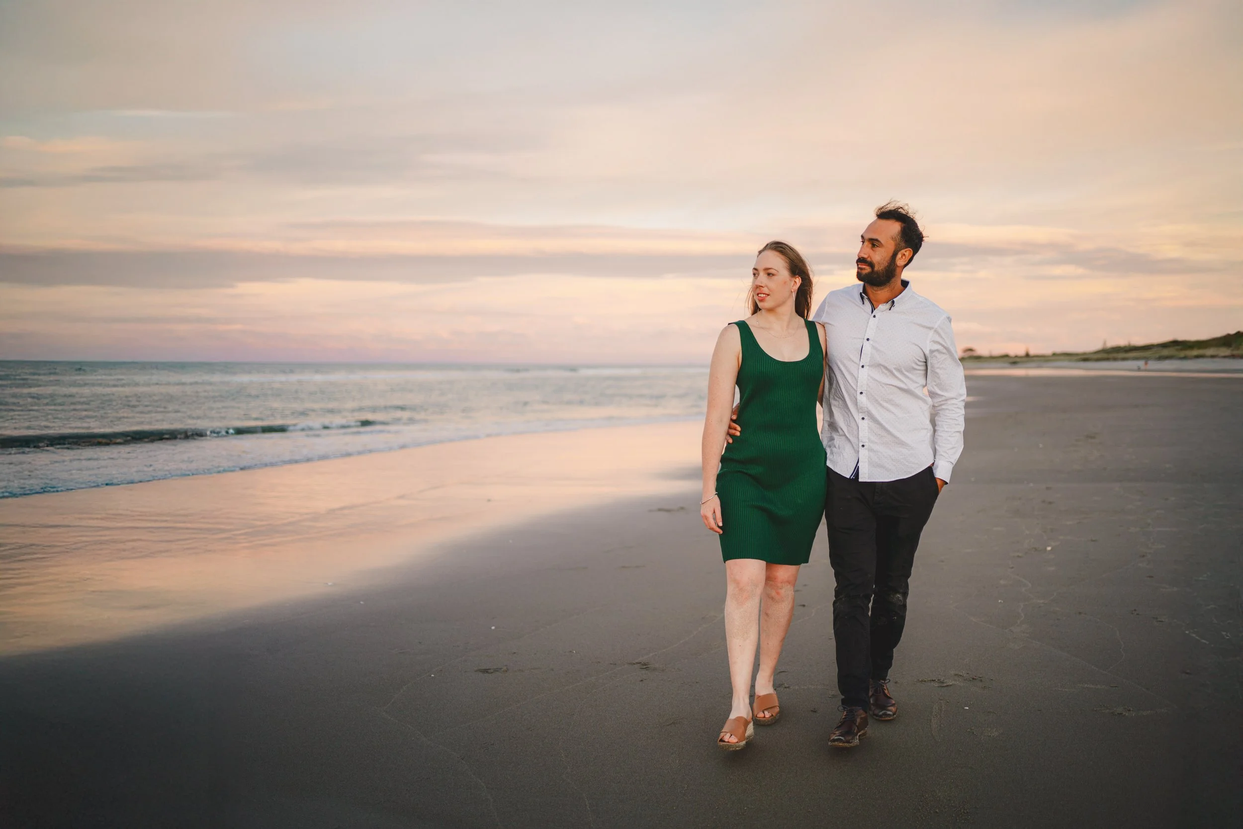 A couple walking along the beach at sunset, with the ocean and sky in the background.