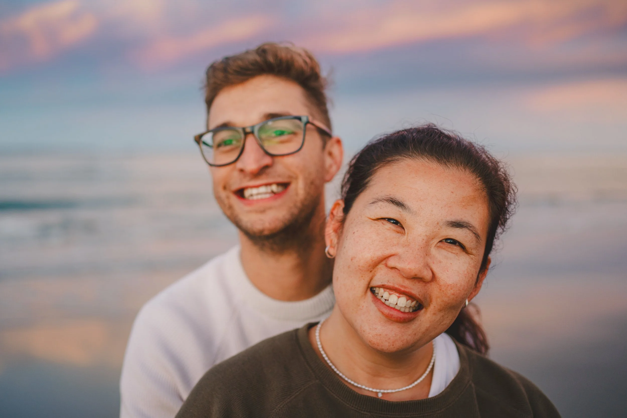 Two people smiling at the camera on a beach at sunset, with colorful sky and reflection in the water behind them.