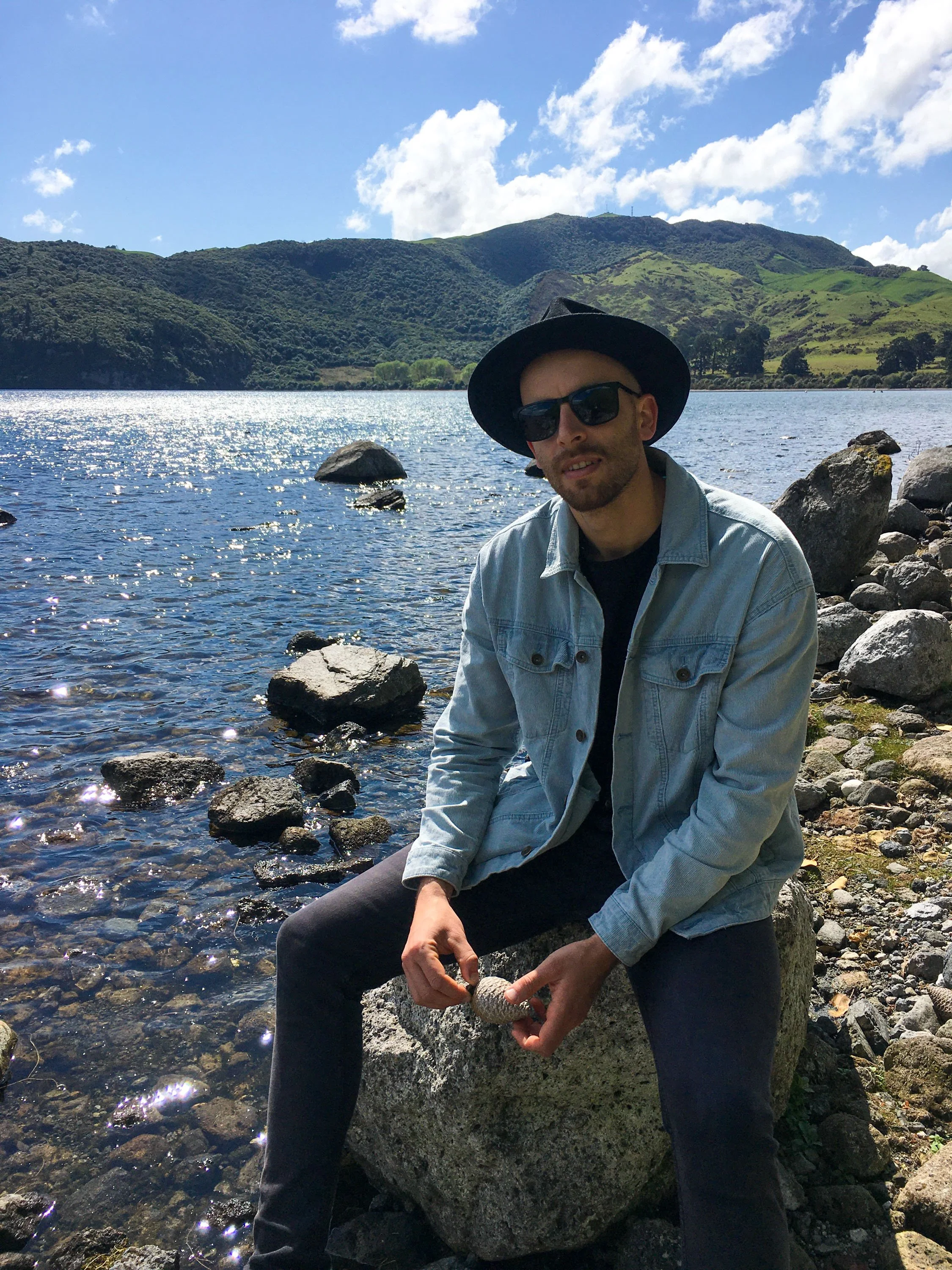 A man sitting on a rock by a lake, wearing a black hat, sunglasses, a light denim jacket, and dark pants, holding a stone, with mountains and a blue sky with clouds in the background.