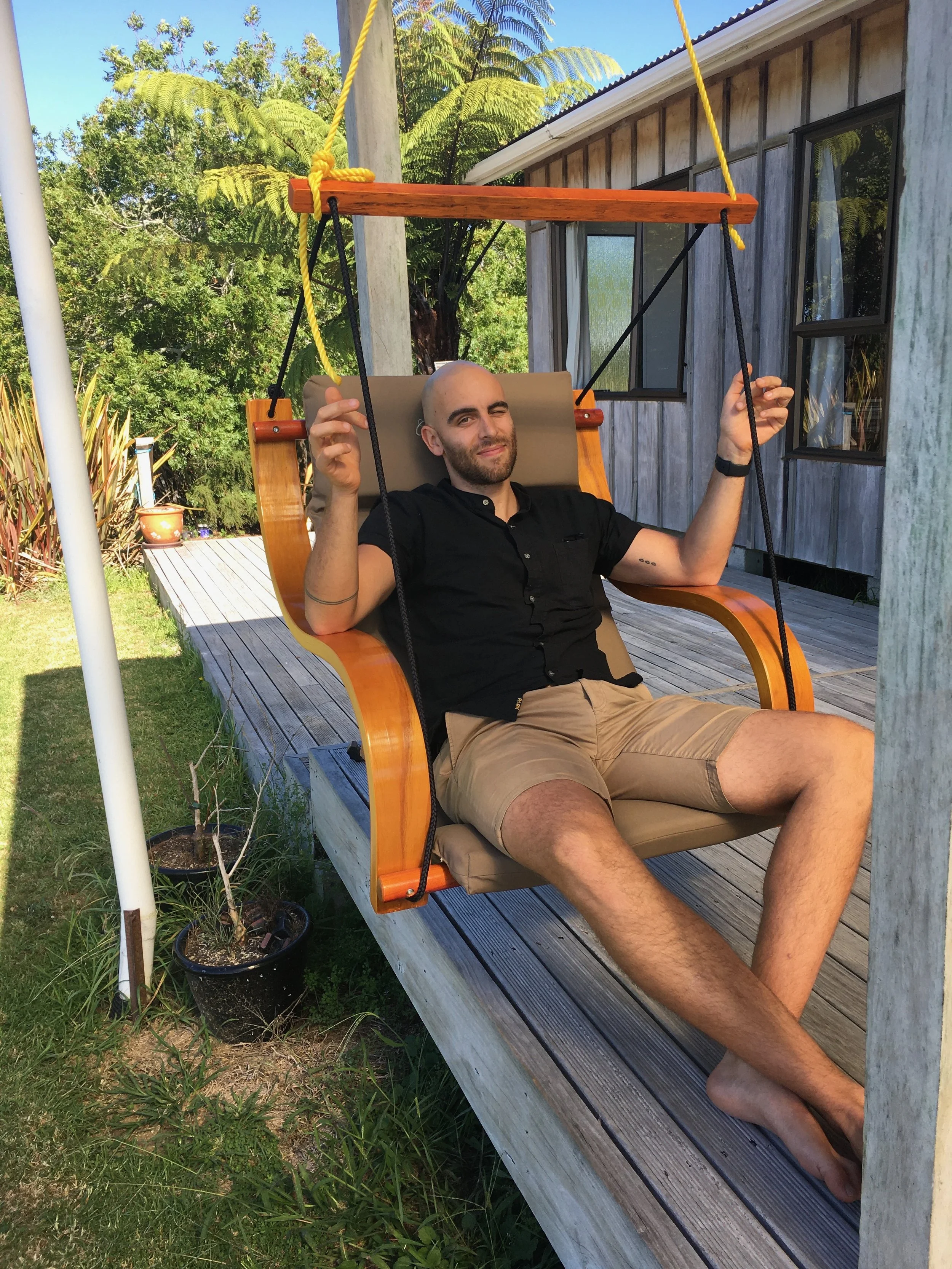A man with a beard and bald head sitting relaxed on an outdoor wooden swing with a cushioned seat, hanging from a wooden frame, on a patio with greenery and a house in the background.