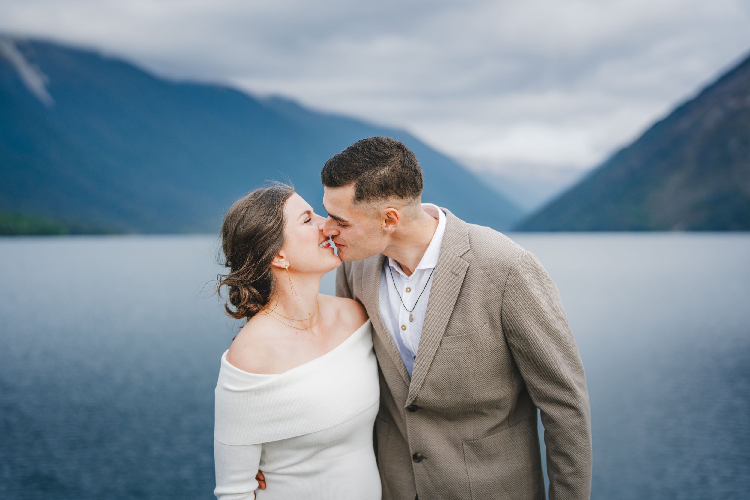A couple kissing outdoors near a lake, with mountains and cloudy sky in the background.
