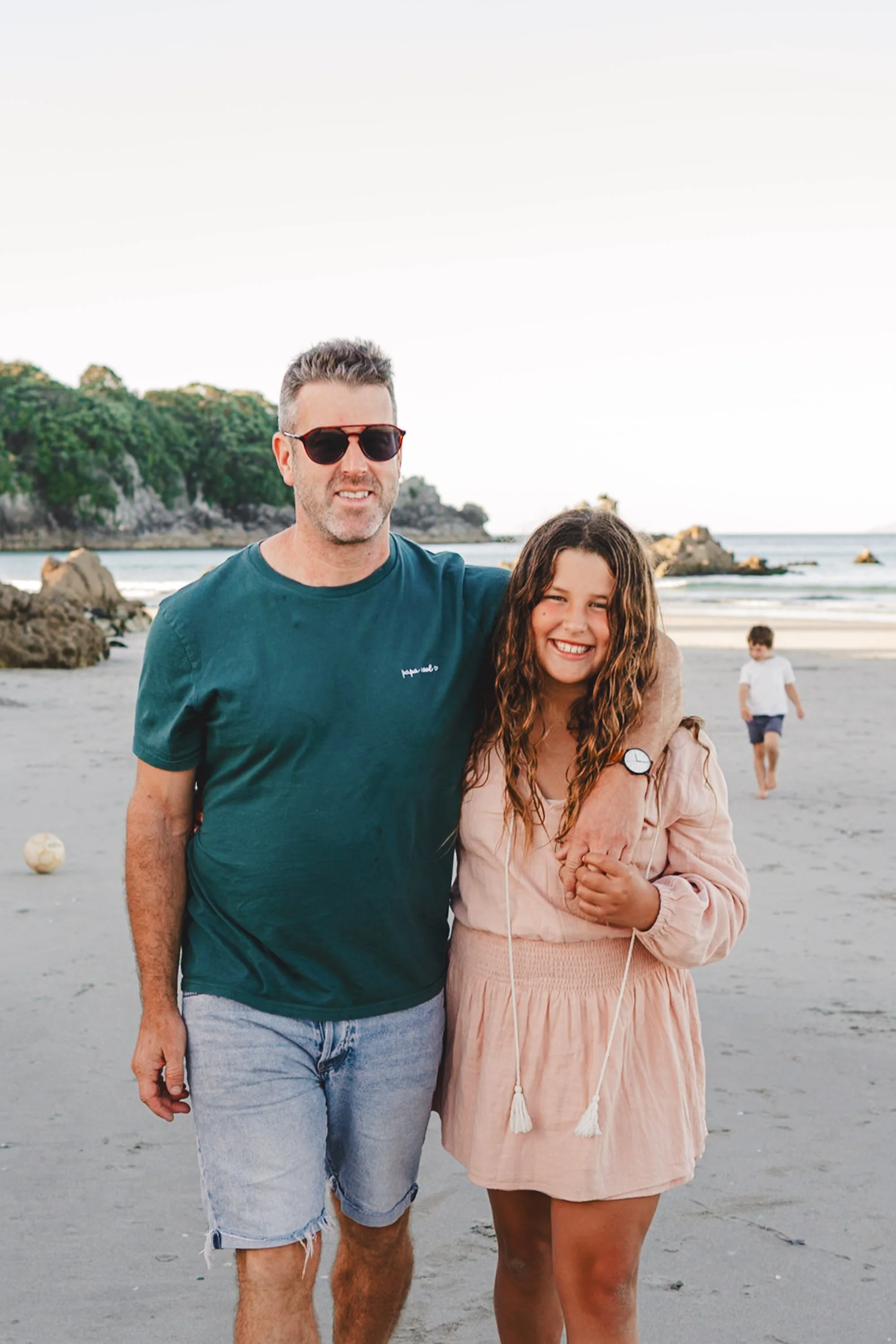 A man and a girl smiling on the beach, with a boy in the background walking along the shoreline, rocky cliffs, and ocean waves.