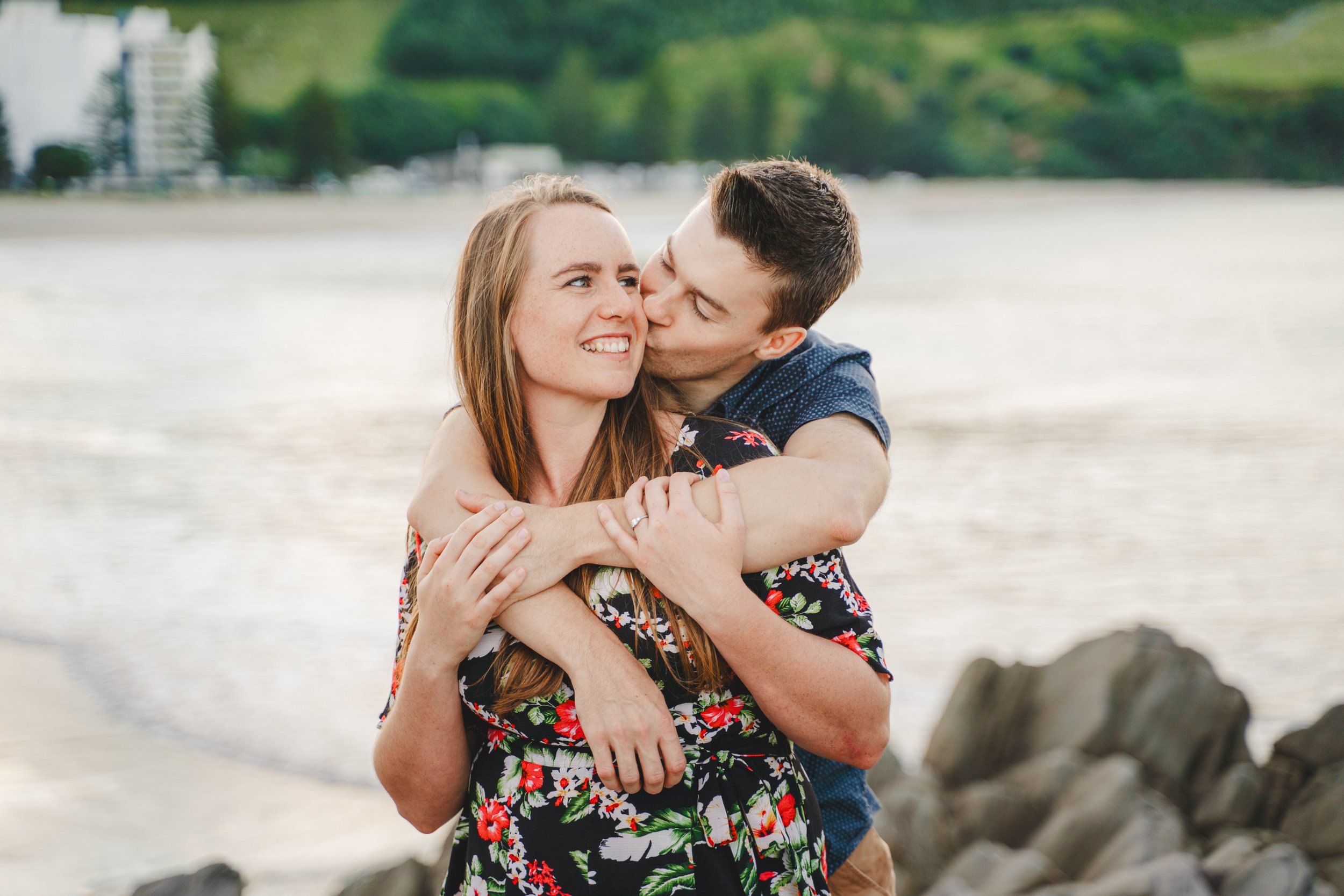A young couple embraces on a rocky beach with a city skyline and green hills in the background. The man is kissing the woman on the side of her face while she smiles.