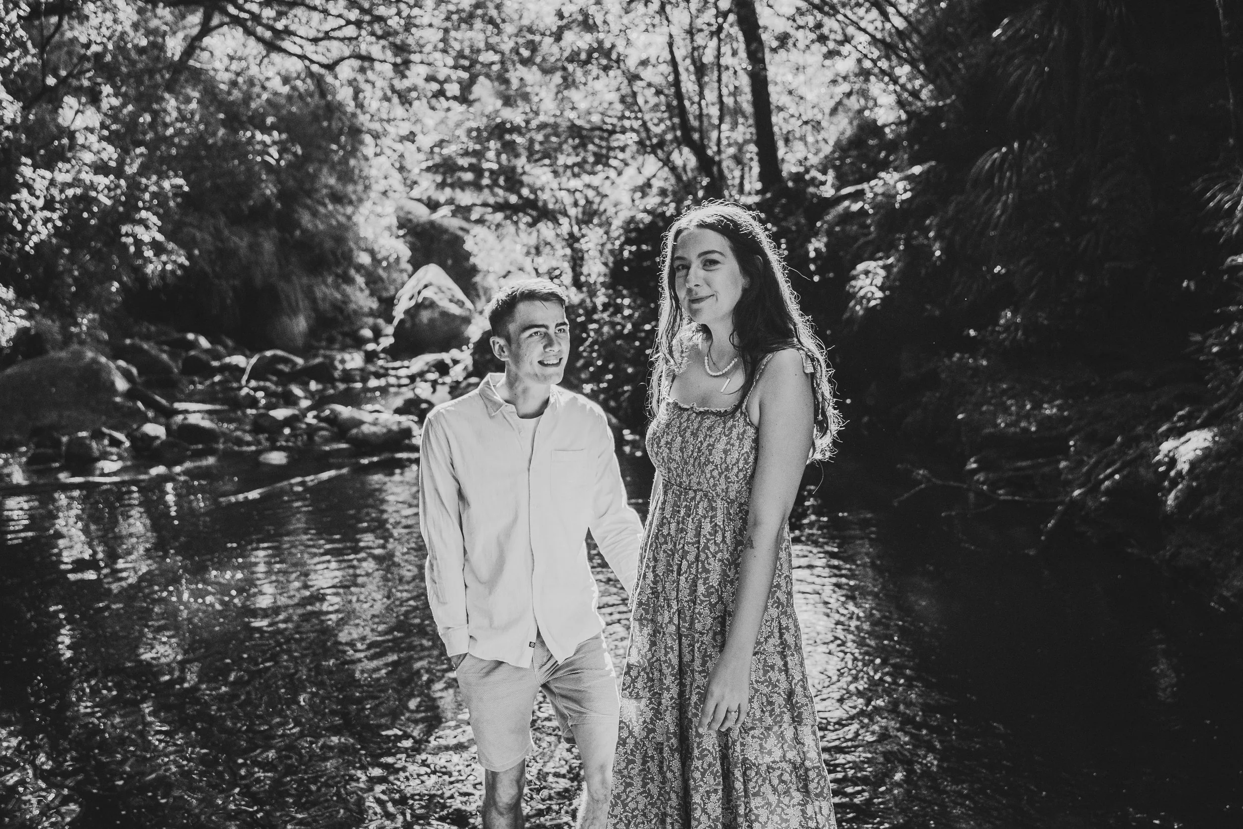 A black and white photo of a young man and woman standing in a shallow stream outdoors, surrounded by trees and rocks, with sunlight filtering through the foliage.