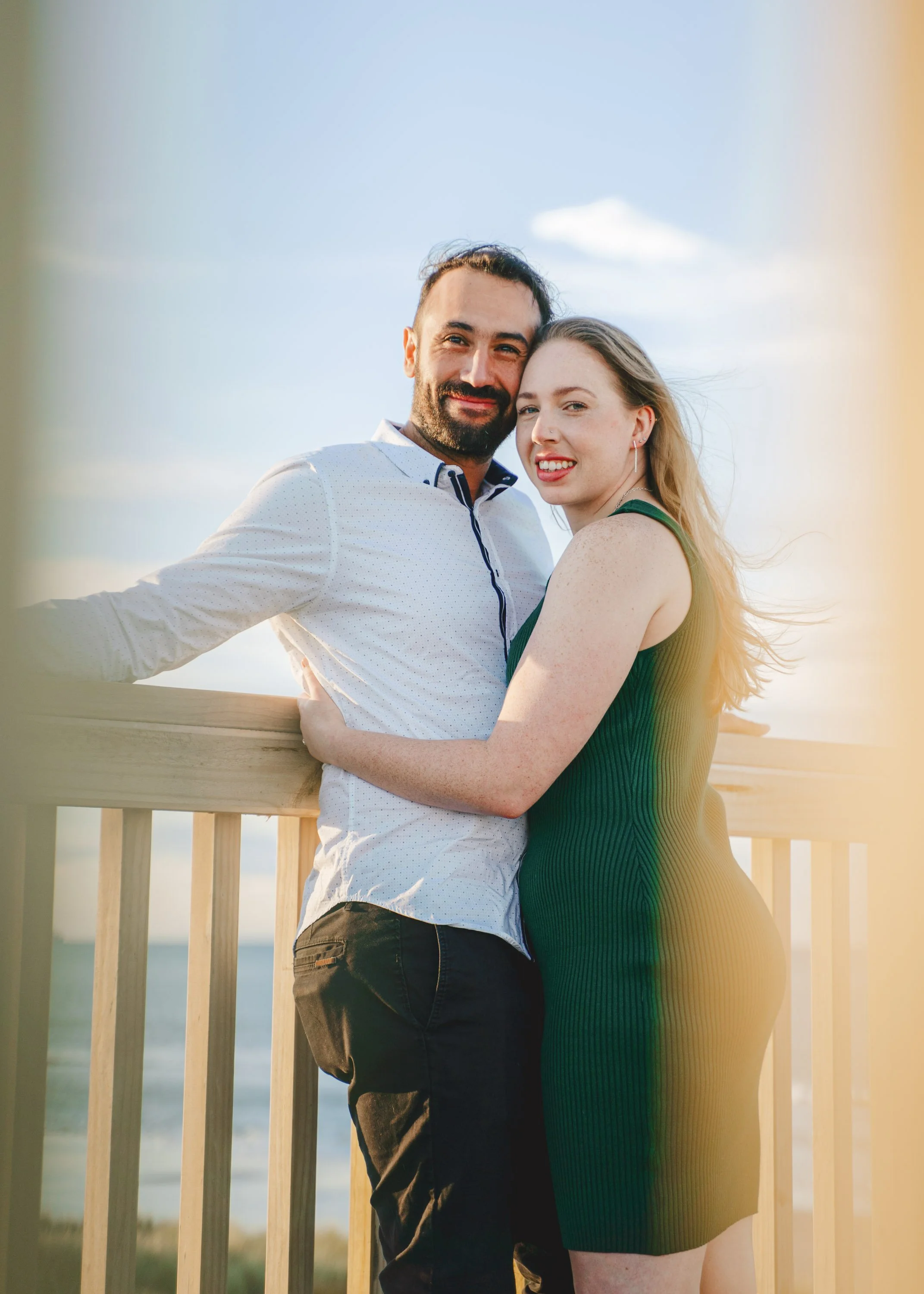 A smiling couple embracing on a wooden deck near the beach at sunset, with the ocean in the background.
