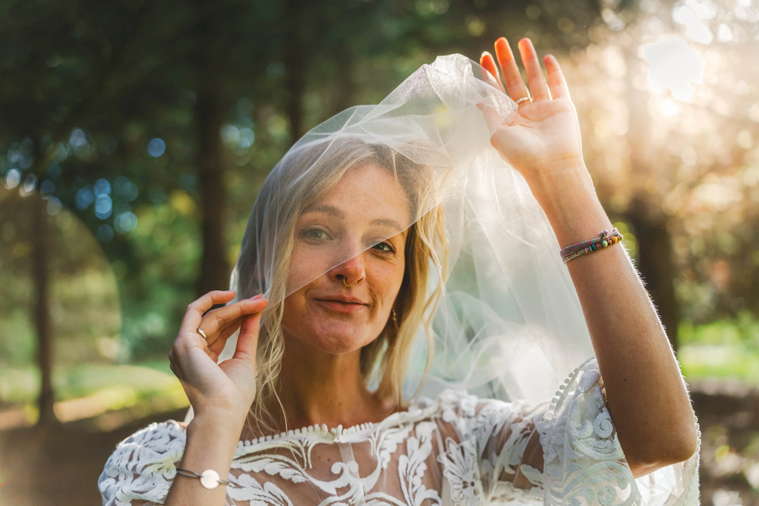 A woman with blonde hair and a nose ring is smiling and holding a veil over her face outdoors with sunlight filtering through trees.