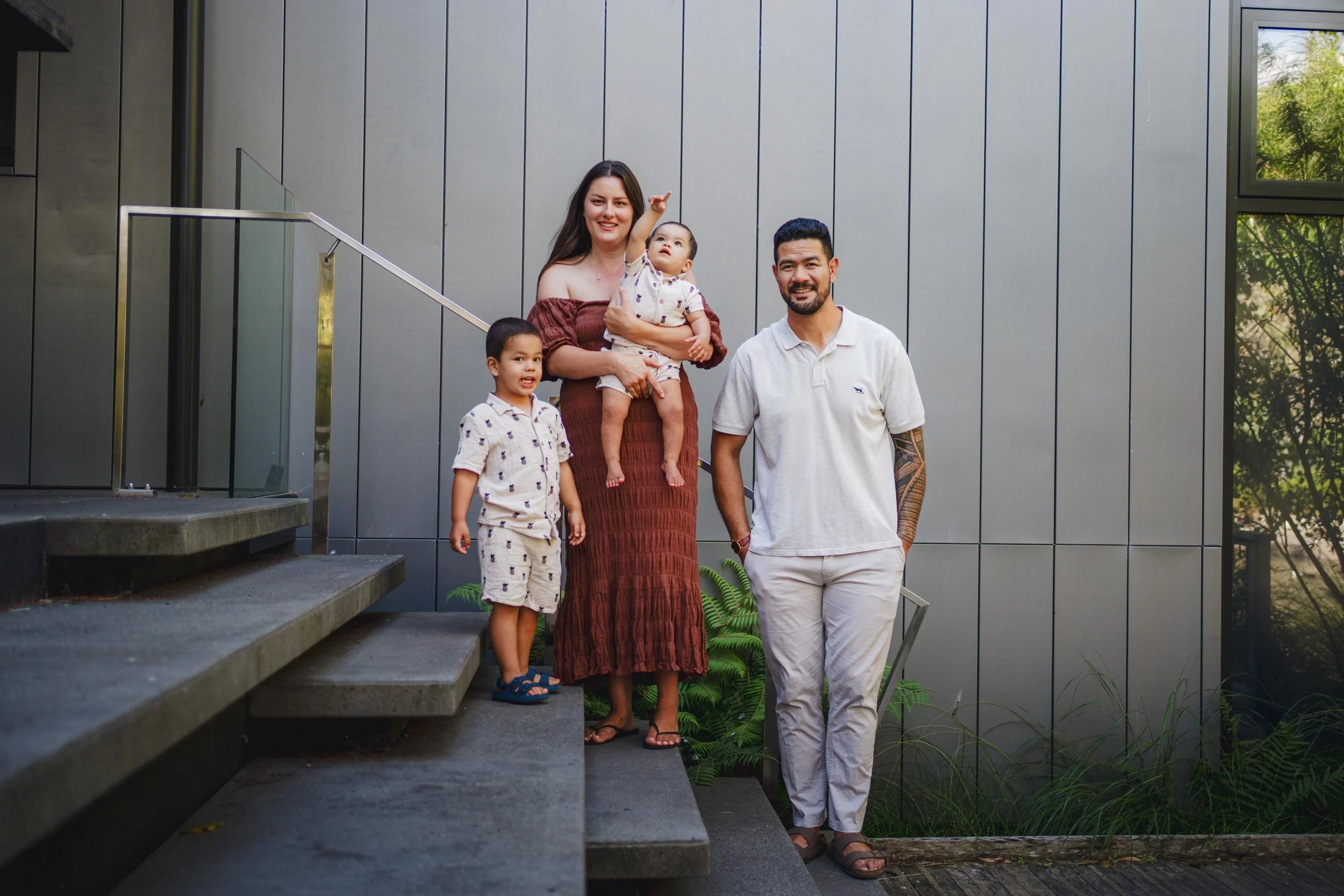 Family of four standing on outdoor stairs in front of a modern building wall, smiling and posing for the camera.