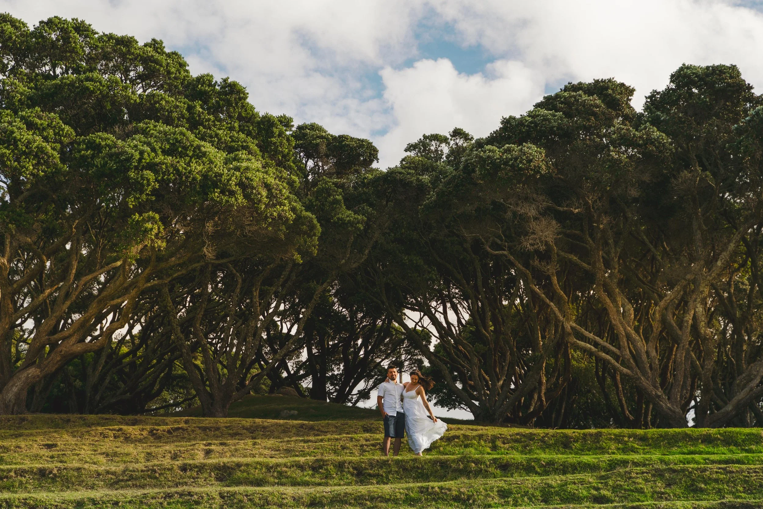 A couple stands on grassy steps under large, lush green trees with a partly cloudy sky overhead.