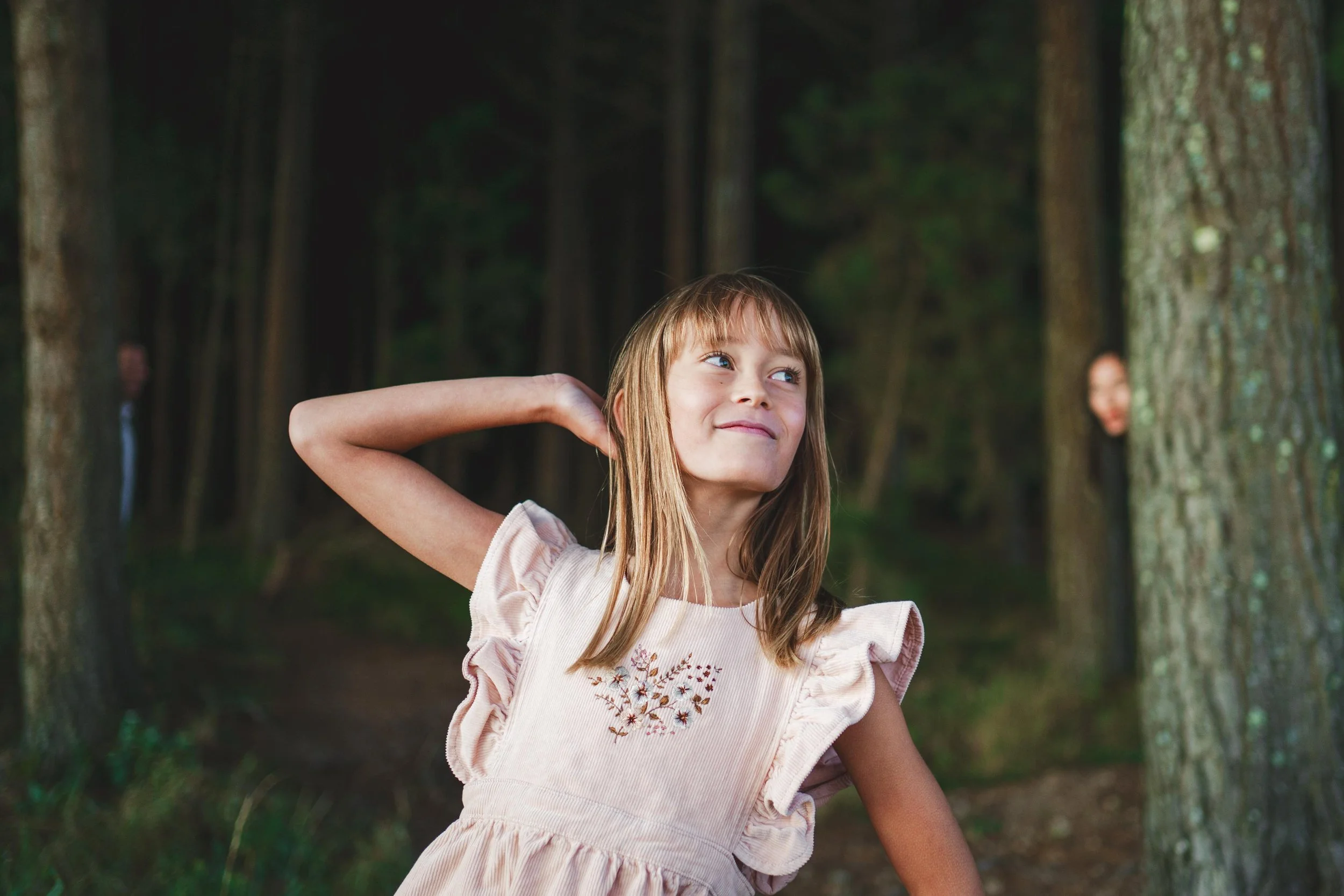 A young girl with light brown hair and a peach-colored dress with floral embroidery, standing in a forest, smiling and touching her neck with one hand.
