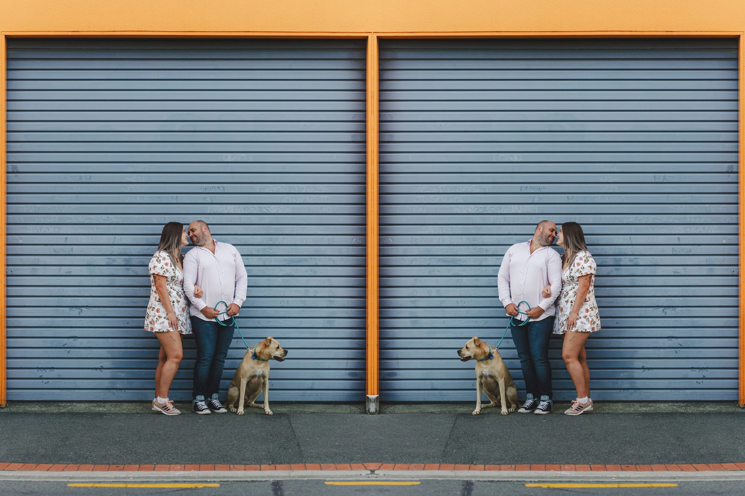 A couple stands with their dog in front of a large, closed, blue metal garage door with orange framing. The woman is wearing a floral dress and the man is in a white shirt and jeans. They are touching foreheads and smiling at each other.