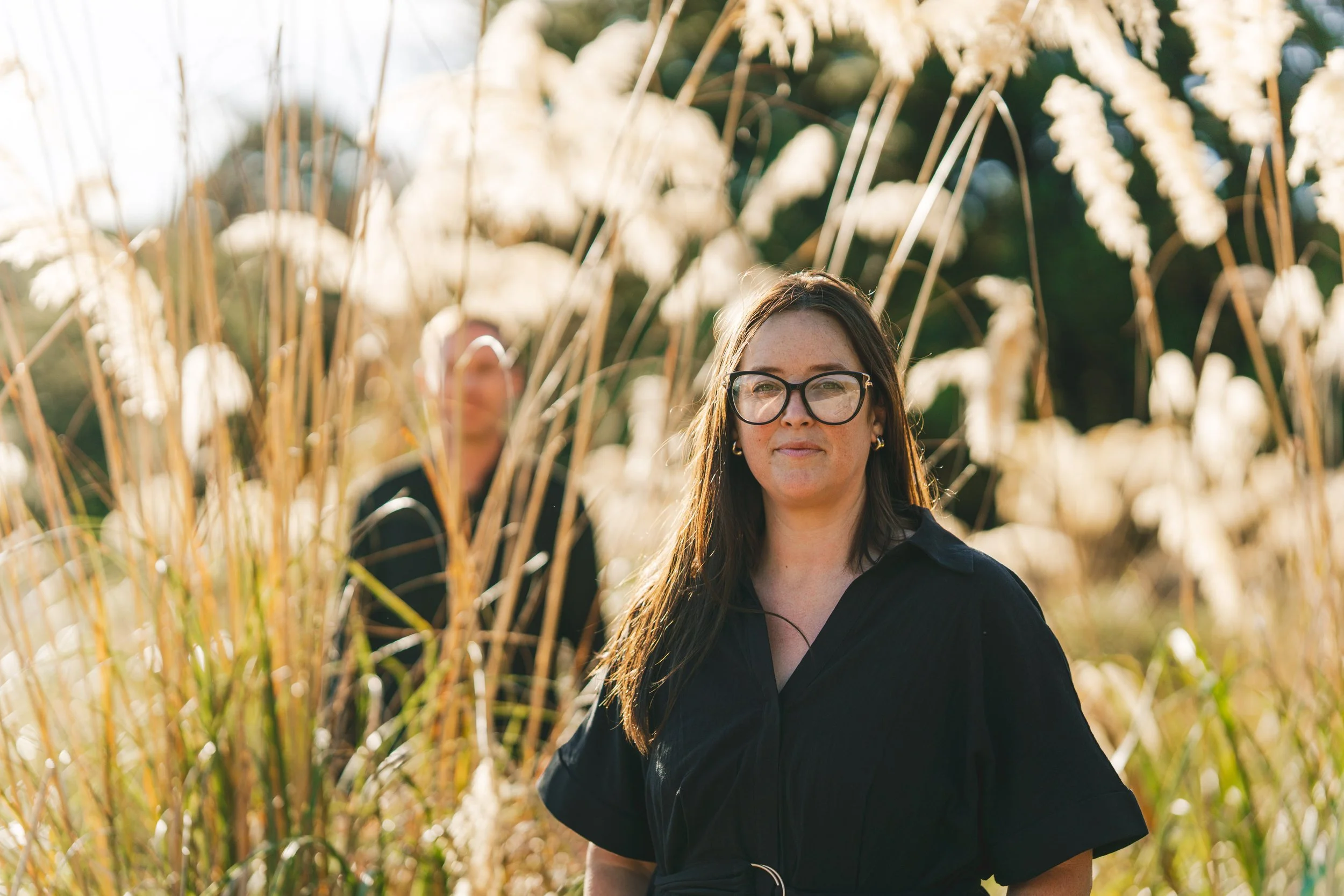 Two women standing in a field of tall, beige grasses during sunset, with one in the foreground wearing glasses and a black shirt, and the other in the background out of focus.