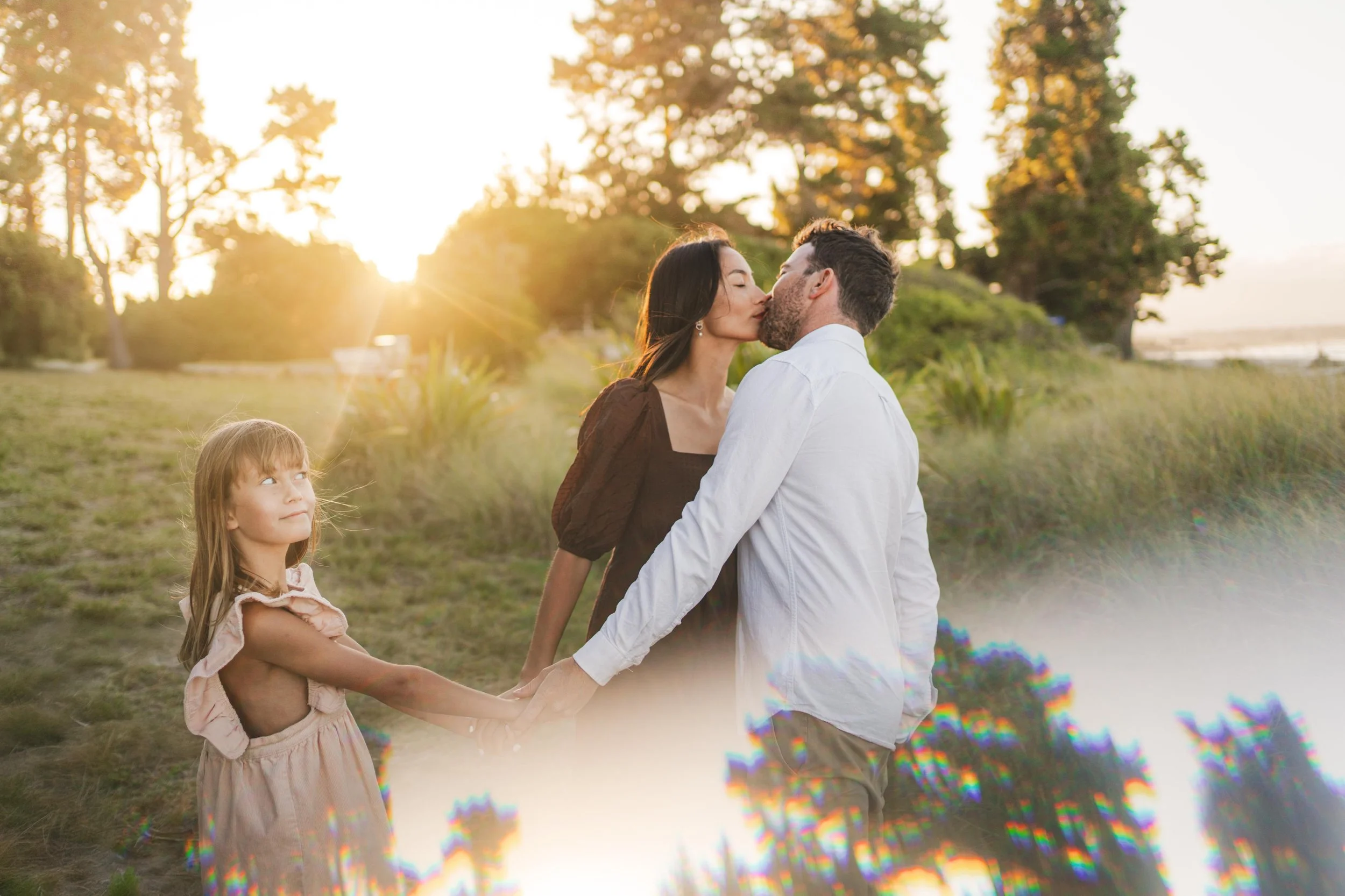 A family outdoor scene at sunset with a man and woman kissing while holding hands, and a young girl standing nearby looking up, in a grassy area with trees in the background.