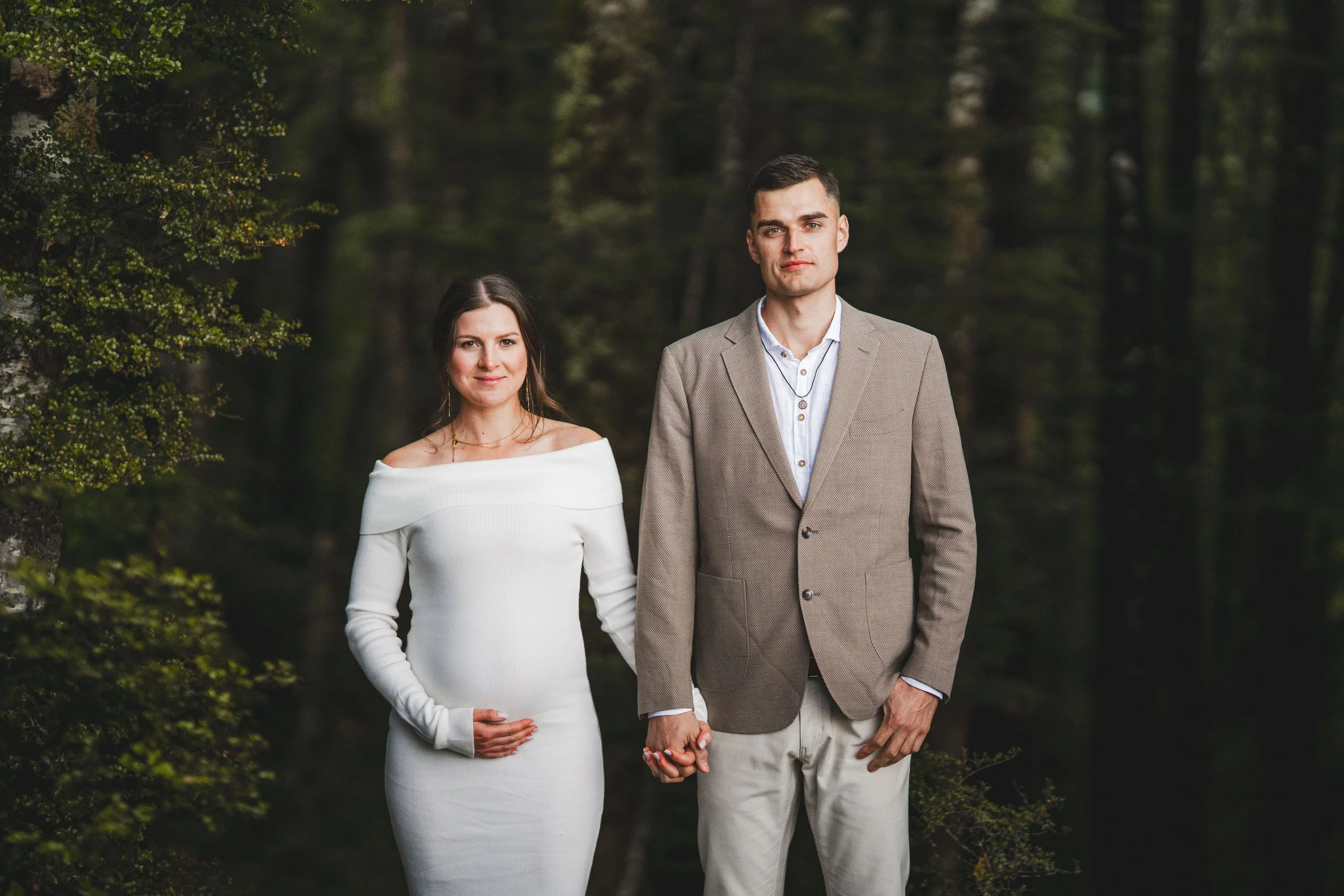 A couple holding hands in a forested area. The woman is wearing a white off-shoulder dress, and the man is in a beige blazer with a white shirt.