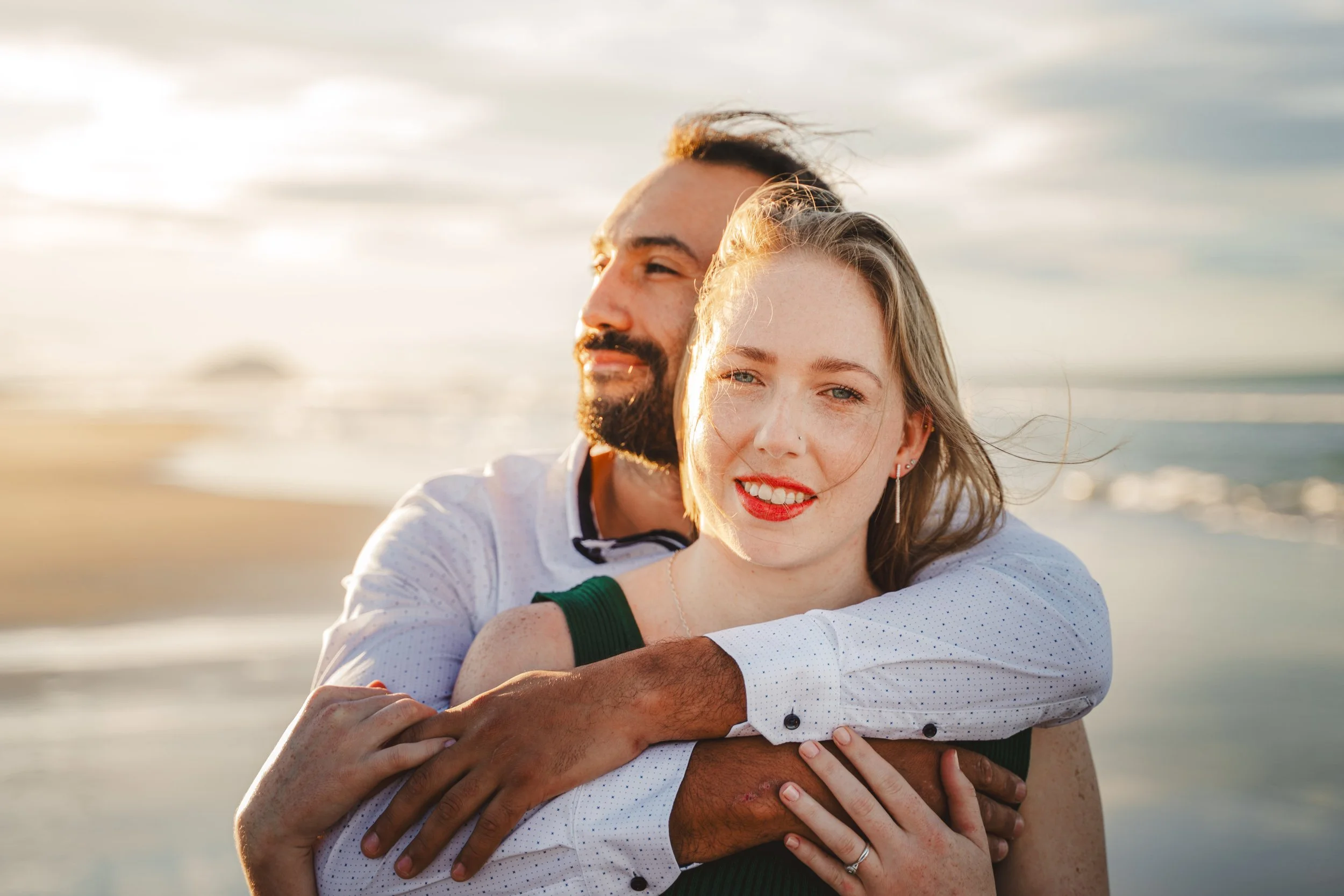 A happy couple embracing on a beach at sunset, with the woman smiling and the man smiling slightly, both facing the camera.