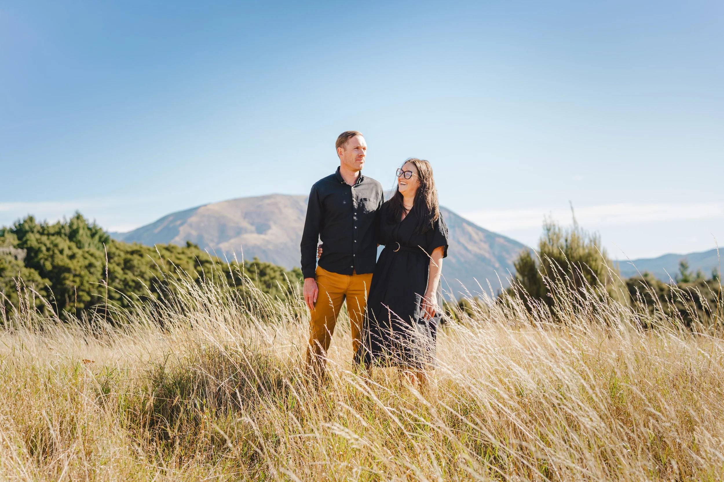 A couple standing in a grassy field with mountains in the background, the man with his arm around the woman who is smiling and wearing glasses.