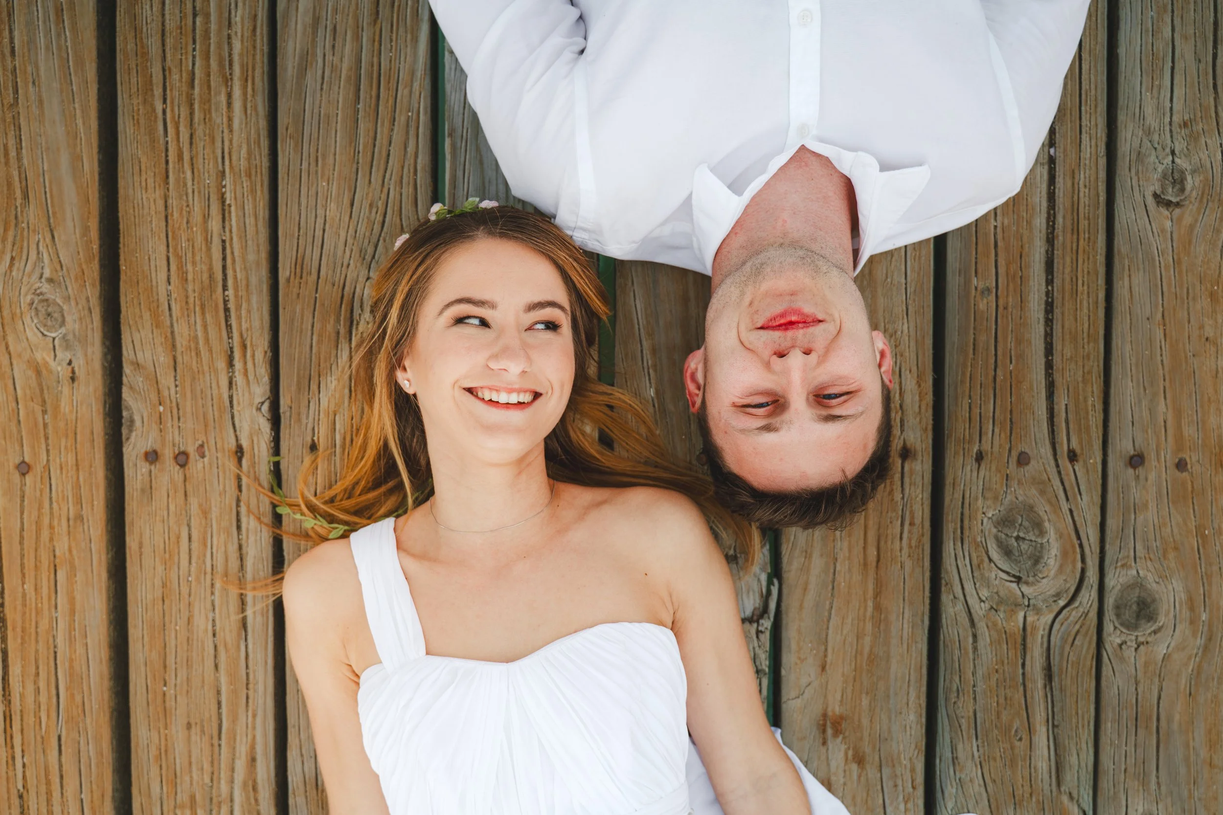 A young woman with long red hair and a white dress is lying on a wooden dock, smiling and looking at a man with short dark hair and a white shirt who is also lying on the dock, with their heads close together.