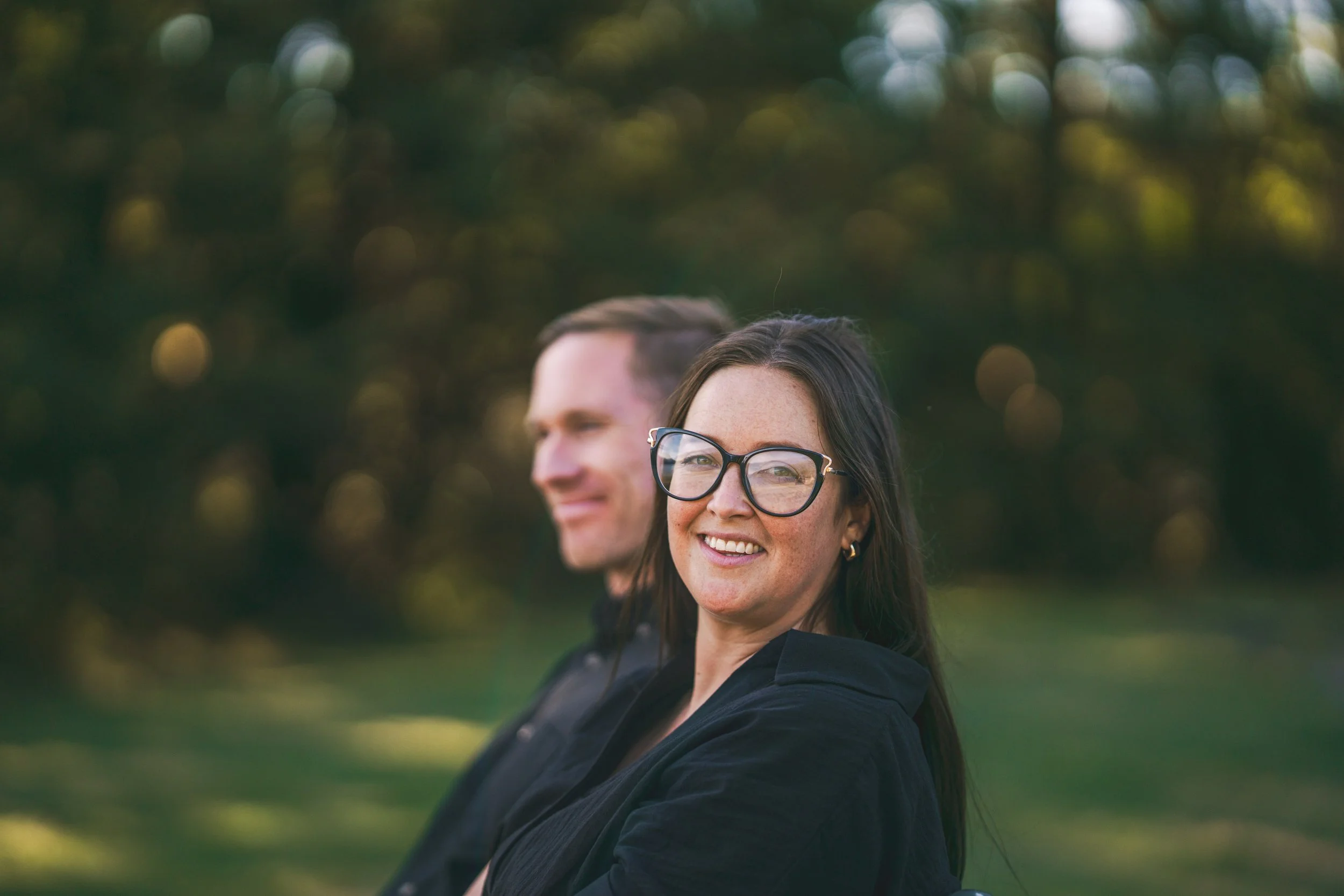A woman with long dark hair, wearing glasses, smiling at the camera, and a man with short hair smiling in the background, outdoors in a park with blurred green trees.
