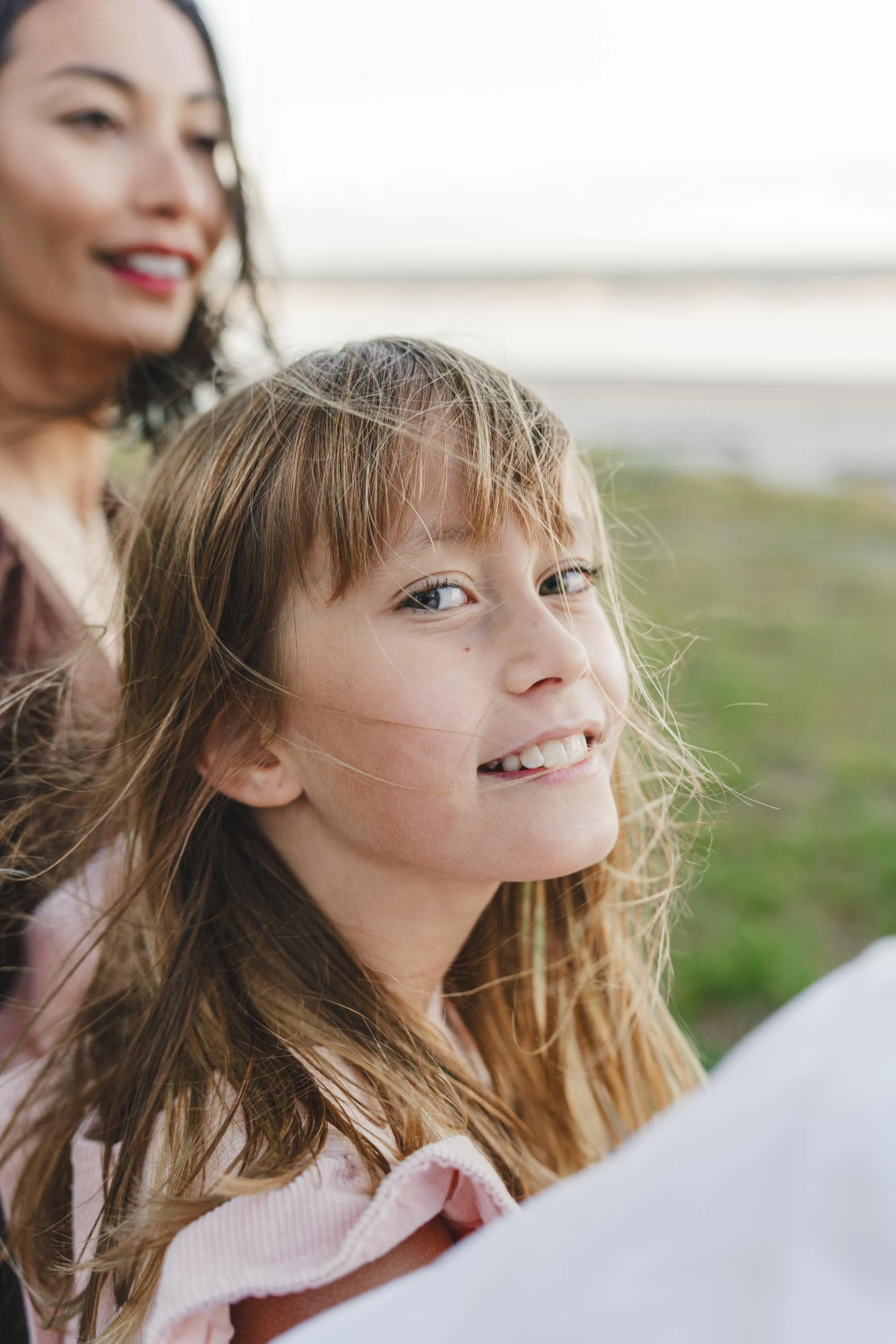 A smiling young girl with red hair and blue eyes outdoors, with an adult woman blurred in the background.