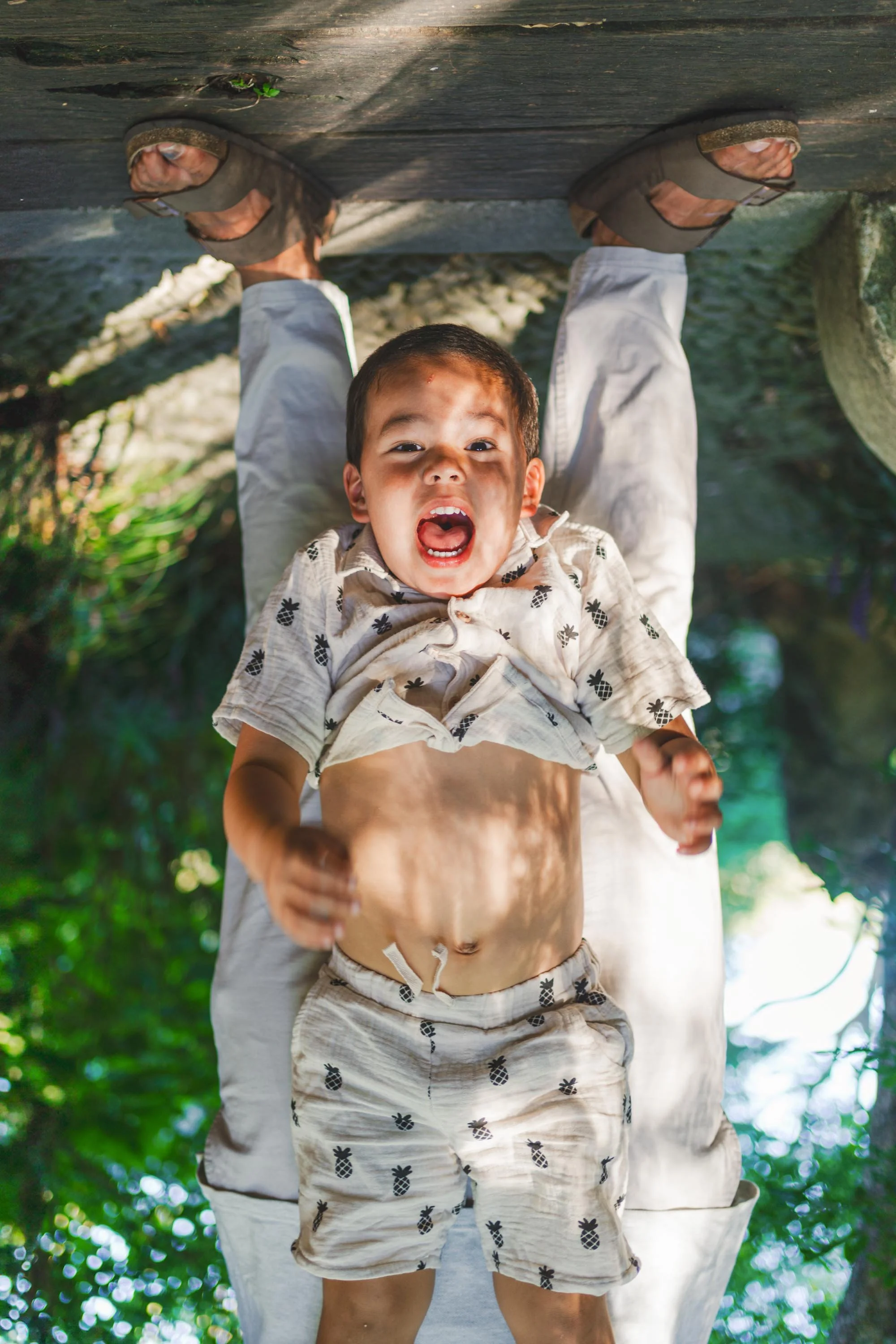 A young boy lying on his back outdoors, making a surprised or excited face, with lush green foliage in the background.