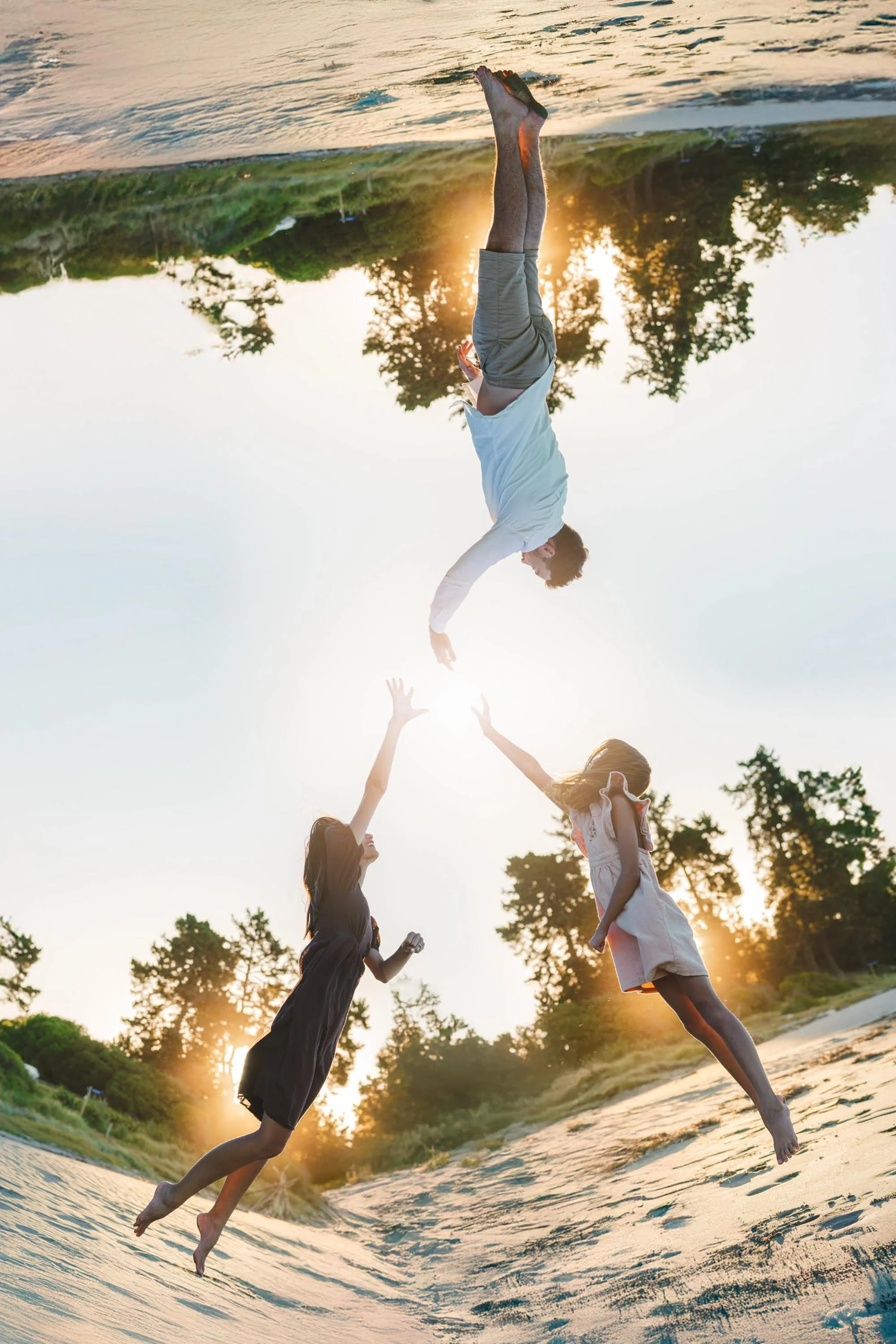 A man and two children, a girl and a boy, are playing and reaching out to each other on a sandy beach during sunset, with trees in the background.