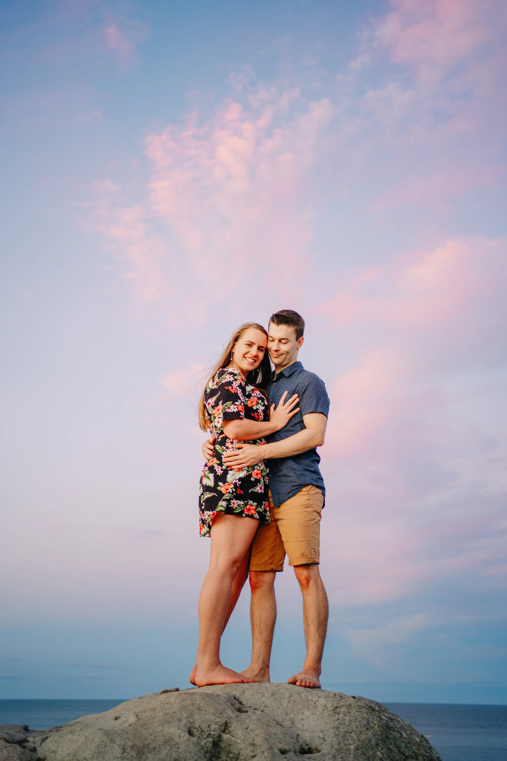 A happy couple embraces on a large rock at the beach during sunset, with a pink and blue sky in the background.