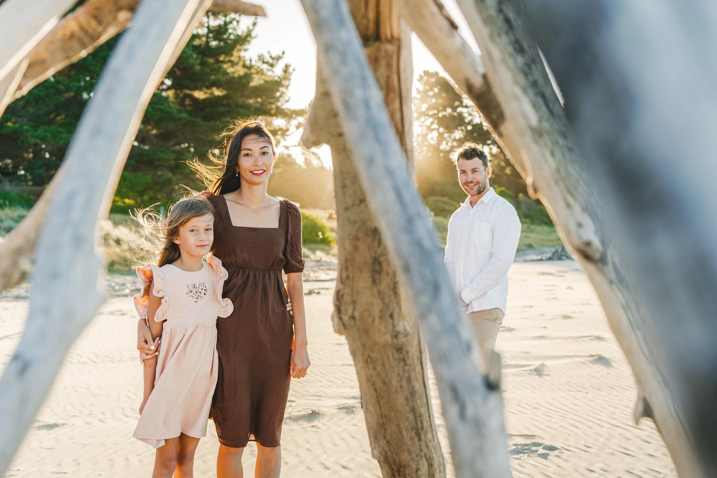 A family of four standing on a beach during sunset, with driftwood in the foreground. The mother and daughter are in the front, and the father is in the background smiling. Trees and sunlight are visible behind them.