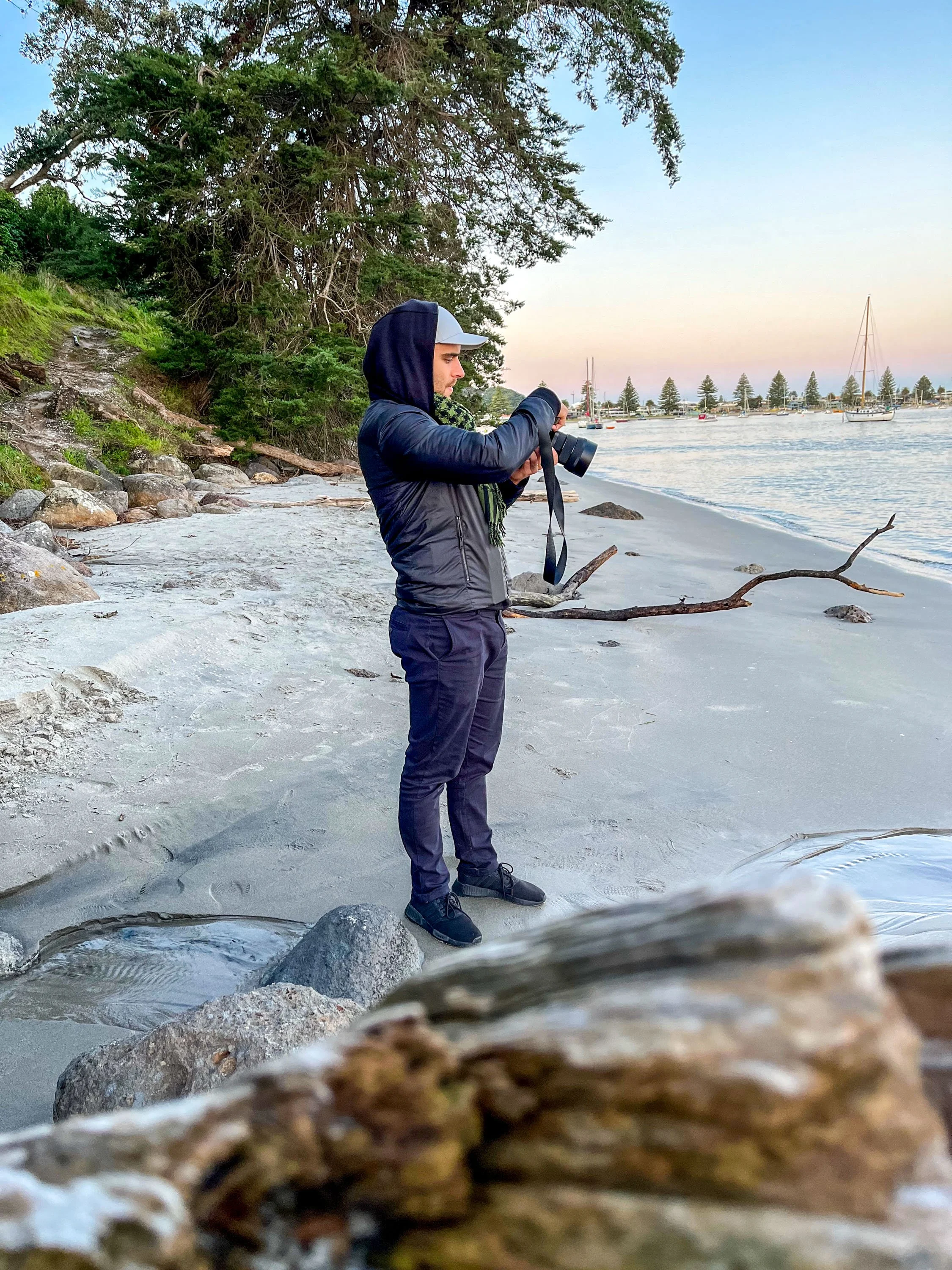 Person standing on a sandy beach near rocks, taking photos with a camera, with boats in the water and trees in the background.