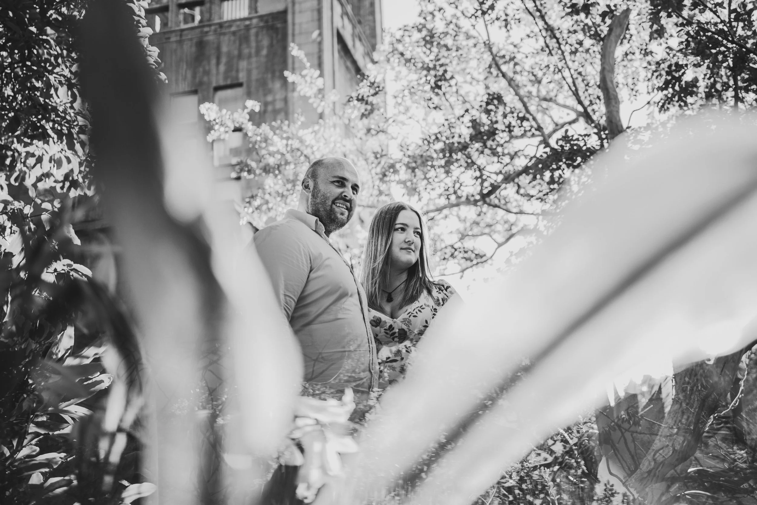 Two people, a man and a woman, standing outdoors surrounded by trees and plants, smiling and looking at something off in the distance.