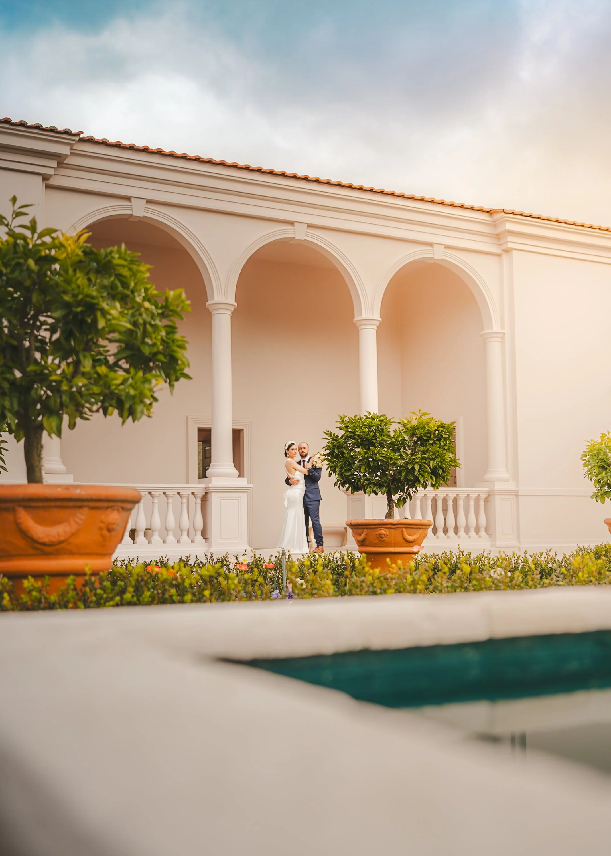 A bride and groom standing together on a balcony with large white columns, surrounded by potted trees and greenery, under a cloudy sky.