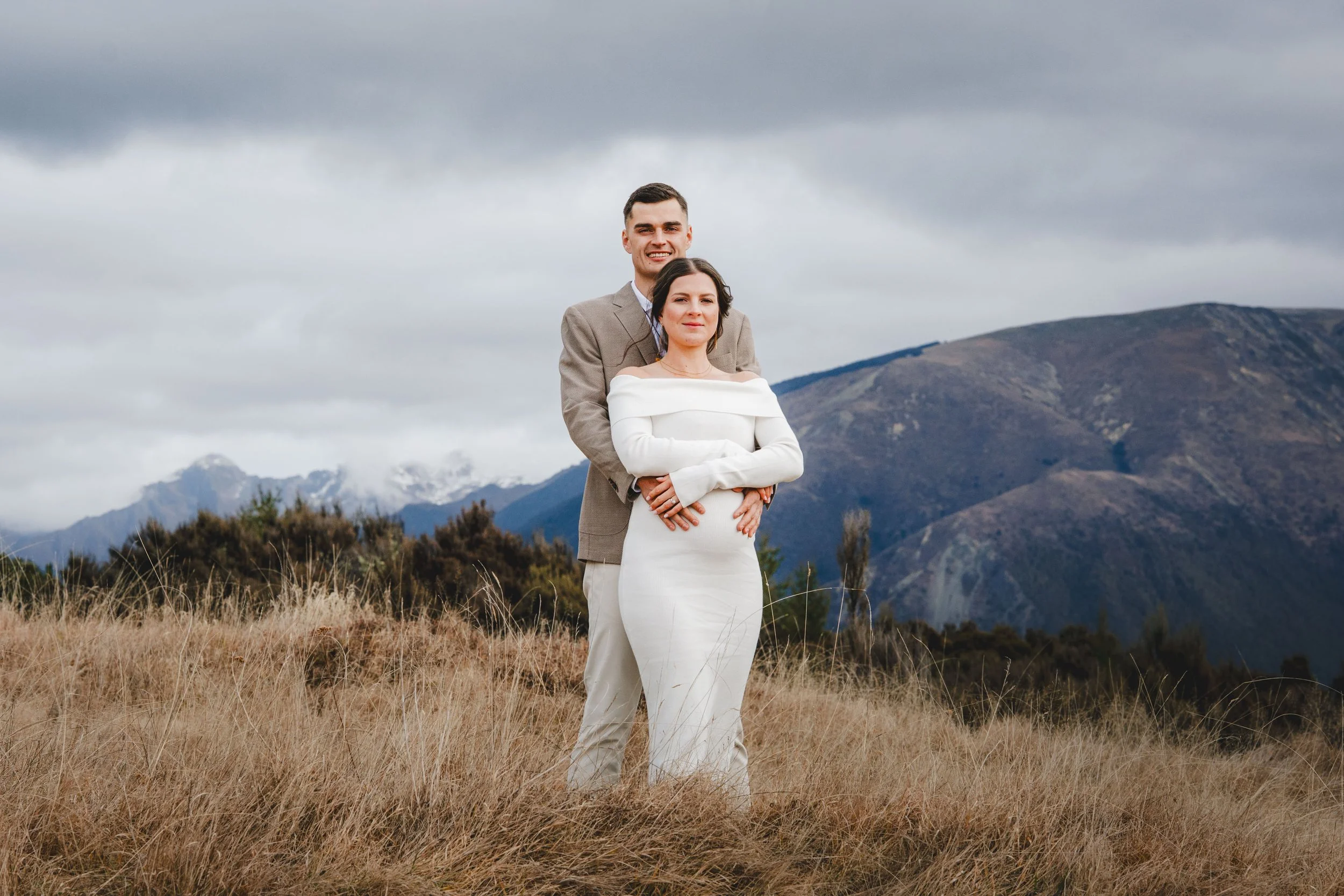 A couple standing in a grassy field with mountains in the background. The woman wears a white off-shoulder dress, and the man wears a gray suit. Both are looking at the camera with neutral expressions.