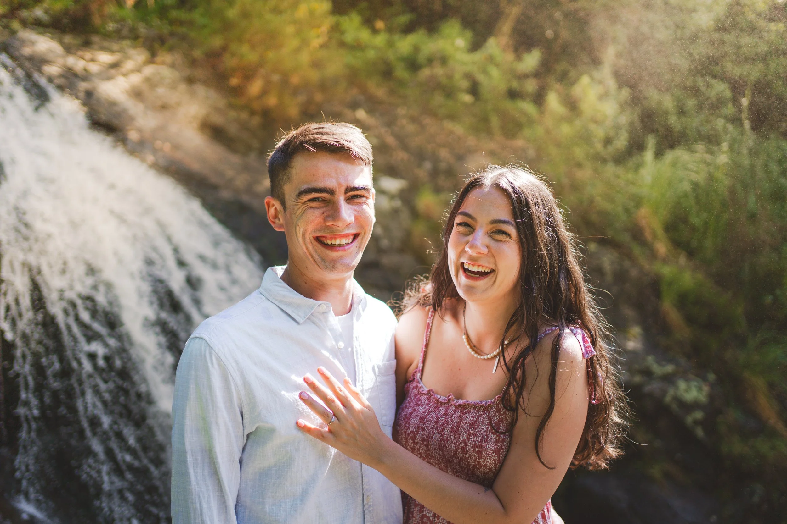 A young couple smiling and laughing outdoors near a waterfall in a forested area, with sunlight and greenery in the background.