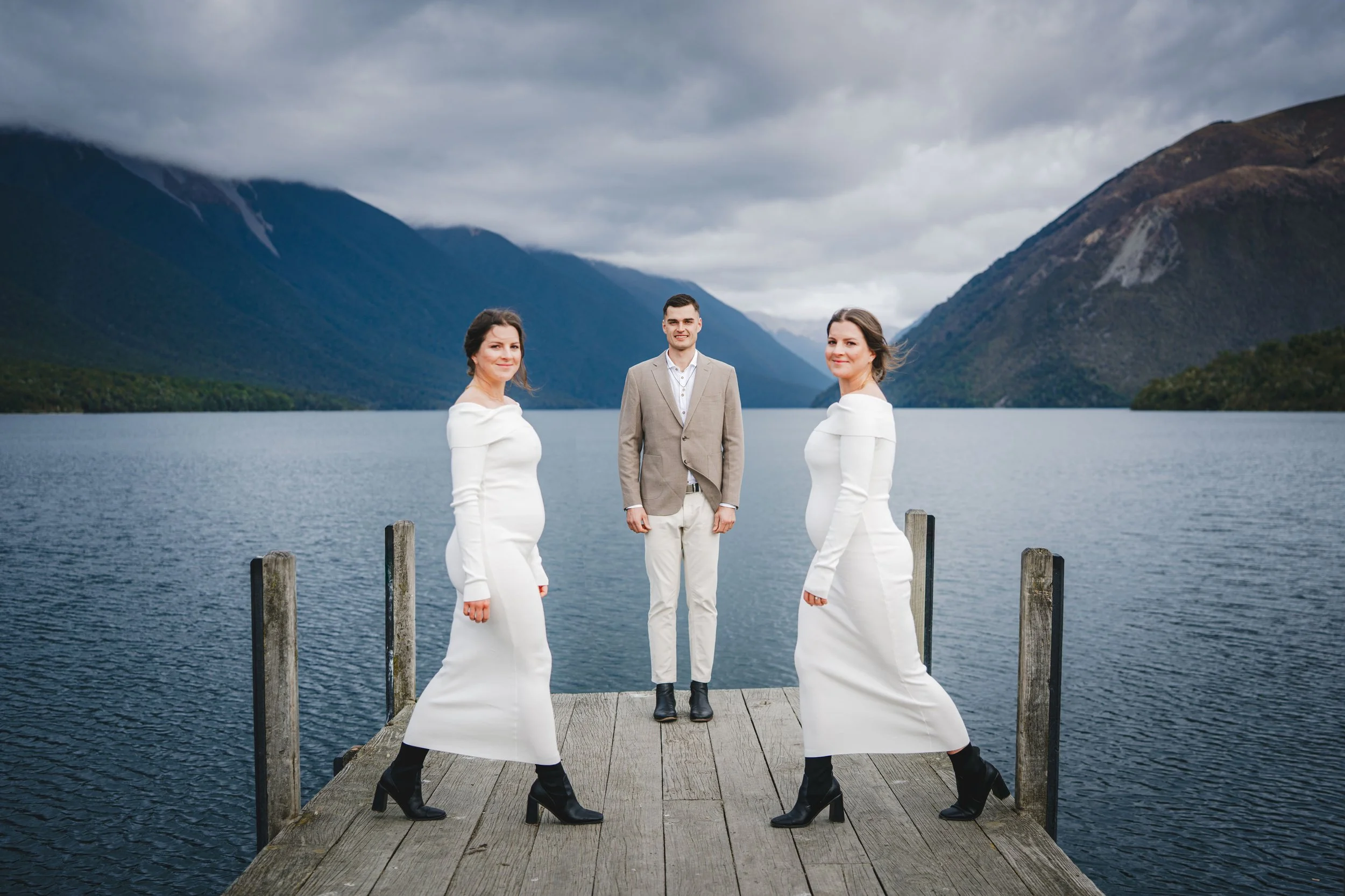 A man and two women standing on a wooden dock by a lake with mountains in the background, dressed in formal attire.